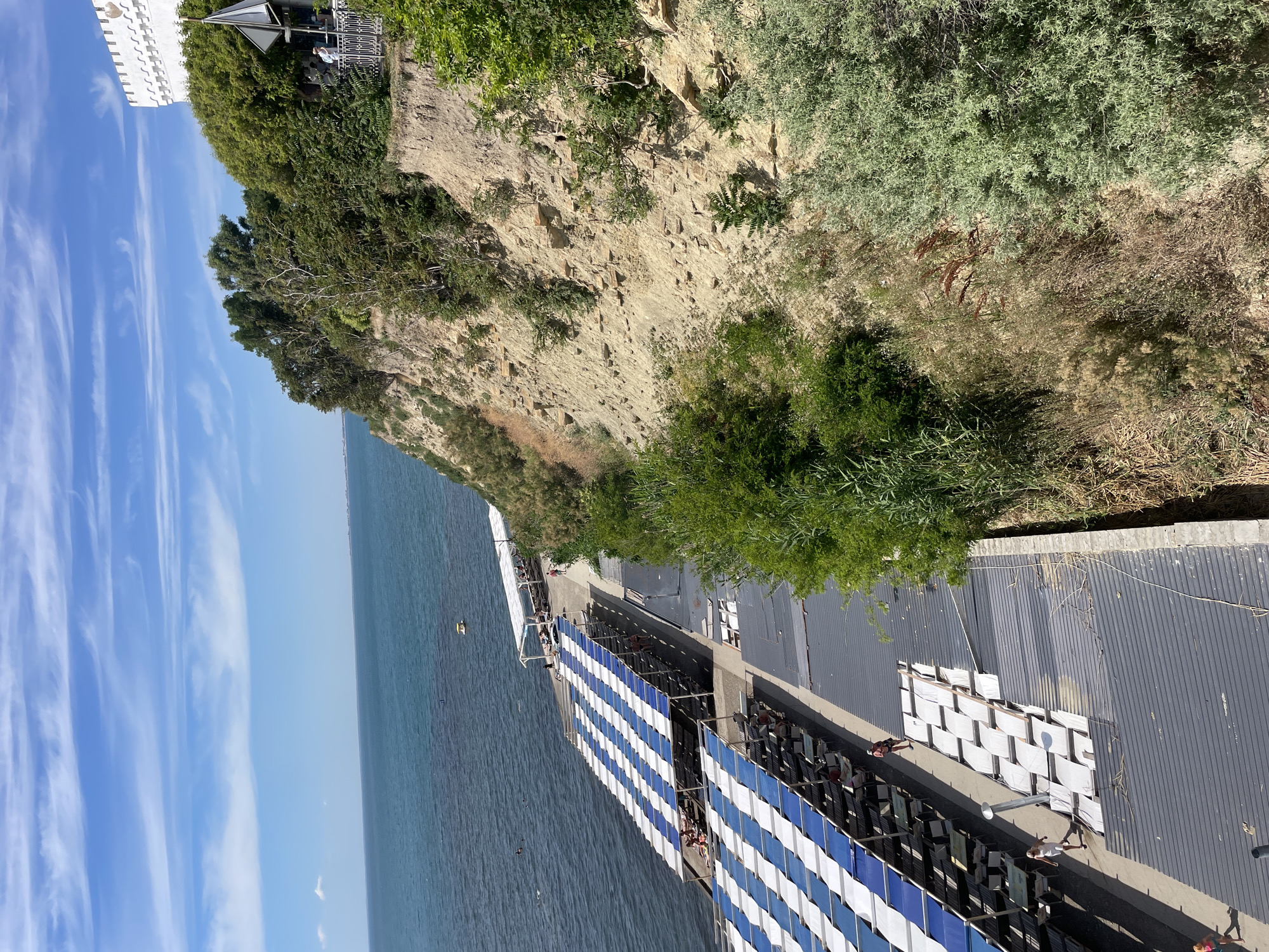 The image depicts an outdoor scene with a cliff overlooking the sea. On the cliff edge, a white building is partially visible, with a structure resembling a gazebo or viewing platform nearby. Lush green vegetation covers the cliff face and the area surrounding the building. Below the cliff, the blue sea stretches to the horizon.
Along the coastline, a man-made structure with rows of seating is set up. These seats are covered with blue and white striped awnings, creating a shaded area. People are seen sitting and standing in this seating area, some appear to be relaxing or watching something. To the right of the seating area, there is a corrugated metal roof structure. The overall setting suggests a public space or venue by the sea, possibly a beach club or event area.
The sky is a clear blue with wispy white clouds, indicating fair weather. The sunlight suggests it is daytime, likely mid-morning or afternoon. The presence of the sea, cliff, and vegetation points to a coastal location. Although the exact event is not clear, the setup with seating implies an organized gathering or a place for spectators. The image has a bright, sunny, and relaxed atmosphere. No visible text is present in the image.