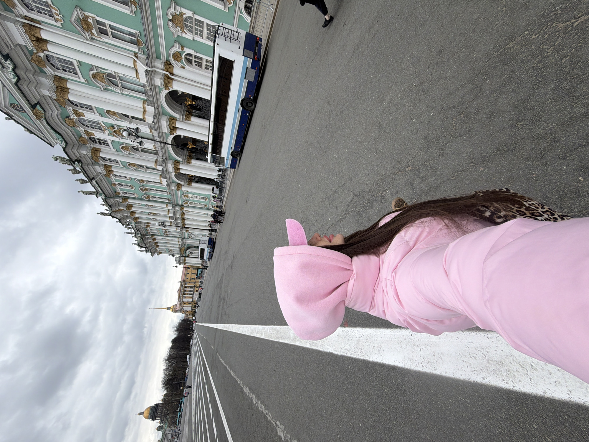 A wide-angle, full-length shot captures a young woman lying on her back on a vast paved square, looking upwards. She wears a soft pink hooded jacket with small ear-like protrusions, and her long dark hair spreads behind her. Her body is positioned along a prominent white line painted on the dark asphalt, which stretches towards the horizon.
In the background, on the left, stands the majestic Winter Palace in Saint Petersburg, Russia, with its distinctive light green facade, numerous white columns, arched windows, and intricate golden architectural details. A blue and white tour bus is parked near the palace. In the far distance, under an expansive, cloudy sky, the golden spire of the Admiralty Building is visible, along with the golden dome of Saint Isaac's Cathedral further to the right. The expansive square is marked with multiple white lines. The overcast sky suggests a cool, daytime setting. The woman's relaxed pose implies she is intentionally striking a unique posture for a photograph in this iconic historic location. No discernible text is visible.
