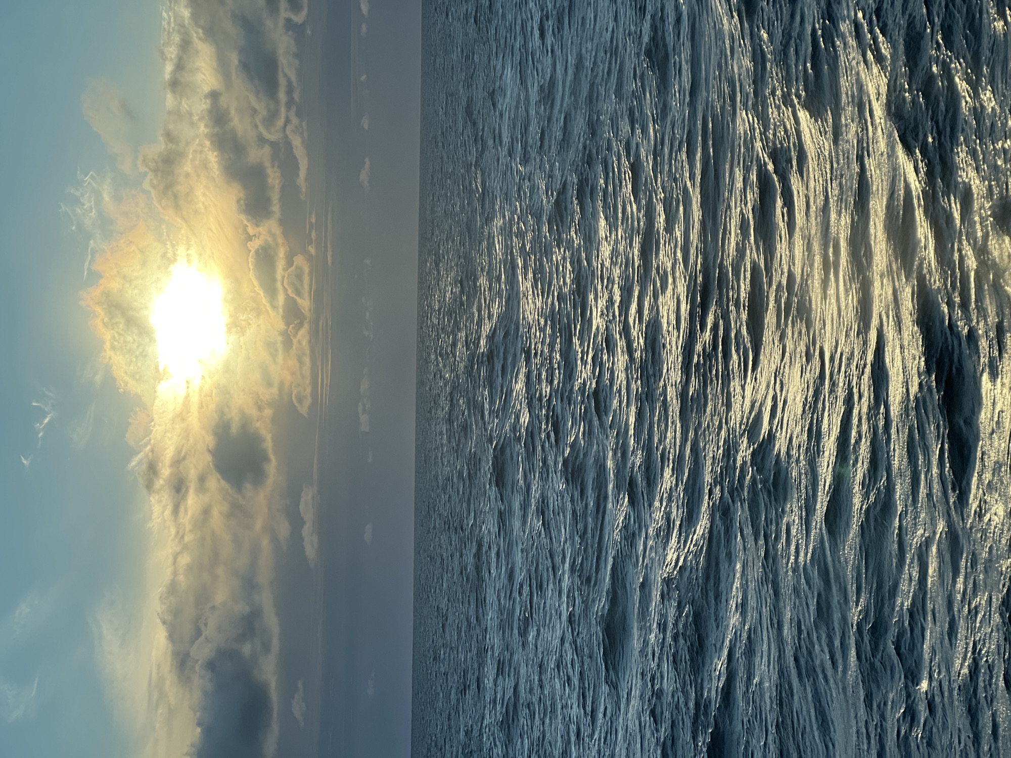 The image depicts a seascape with a prominent sun peeking through scattered clouds, casting a golden reflection on the rippling water. The horizon separates the deep blue ocean from the sky, which transitions from a pale blue at the top to a darker, cloud-laden hue above the water. The waves are small to medium, with the sunlight highlighting the crests of some. There are no people, structures, or specific landmarks visible, rendering it impossible to determine a location within the city of Unknown, Poland, or any other specific geographical area. The environment is solely oceanic.