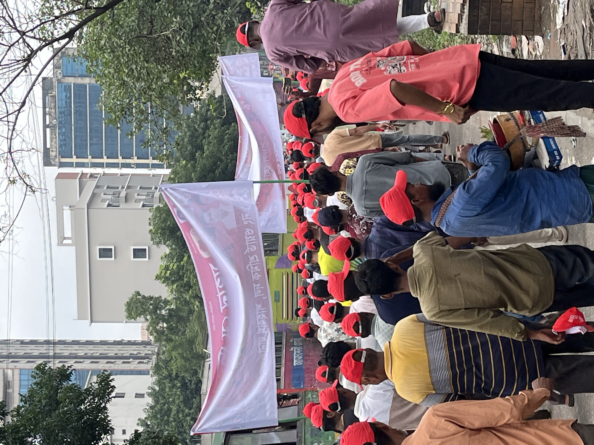 A daytime outdoor scene captures a large gathering of men, predominantly seen from behind or from the side, on what appears to be a street or paved area in an urban environment. Many of the participants are wearing distinctive red caps or hats, suggesting a unified affiliation.
The men are densely packed, stretching into the distance, and appear to be facing forward, possibly towards a speaker or a focal point of the event. Some individuals are standing upright, while others have their heads slightly bowed. In the foreground, one man in a blue shirt is seen holding a small drum or percussion instrument, indicating a possible musical or rhythmic component to the gathering.
Several large white banners with prominent red text in Bengali are visible, erected along the gathering. The text on these banners includes "বাংলাদেশ ছাত্রলীগ" (Bangladesh Chhatra League), which is a major student political organization. Other discernible phrases on the banners relate to political issues, such as "সংবিধান বিরোধী, ভোট ডাকাতি" (anti-constitution, vote rigging) and "শিক্ষা প্রতিষ্ঠানে" (in educational institutions), strongly indicating that this is a political rally or demonstration.
In the background, tall city buildings with visible windows and facades rise above the trees, confirming an urban setting. A large, possibly public transport vehicle (like a bus or truck) in yellow and green is partially visible behind one of the banners. The lighting suggests an overcast day. The overall atmosphere points to an organized public event, likely a political protest or meeting in Dhaka, Bangladesh.