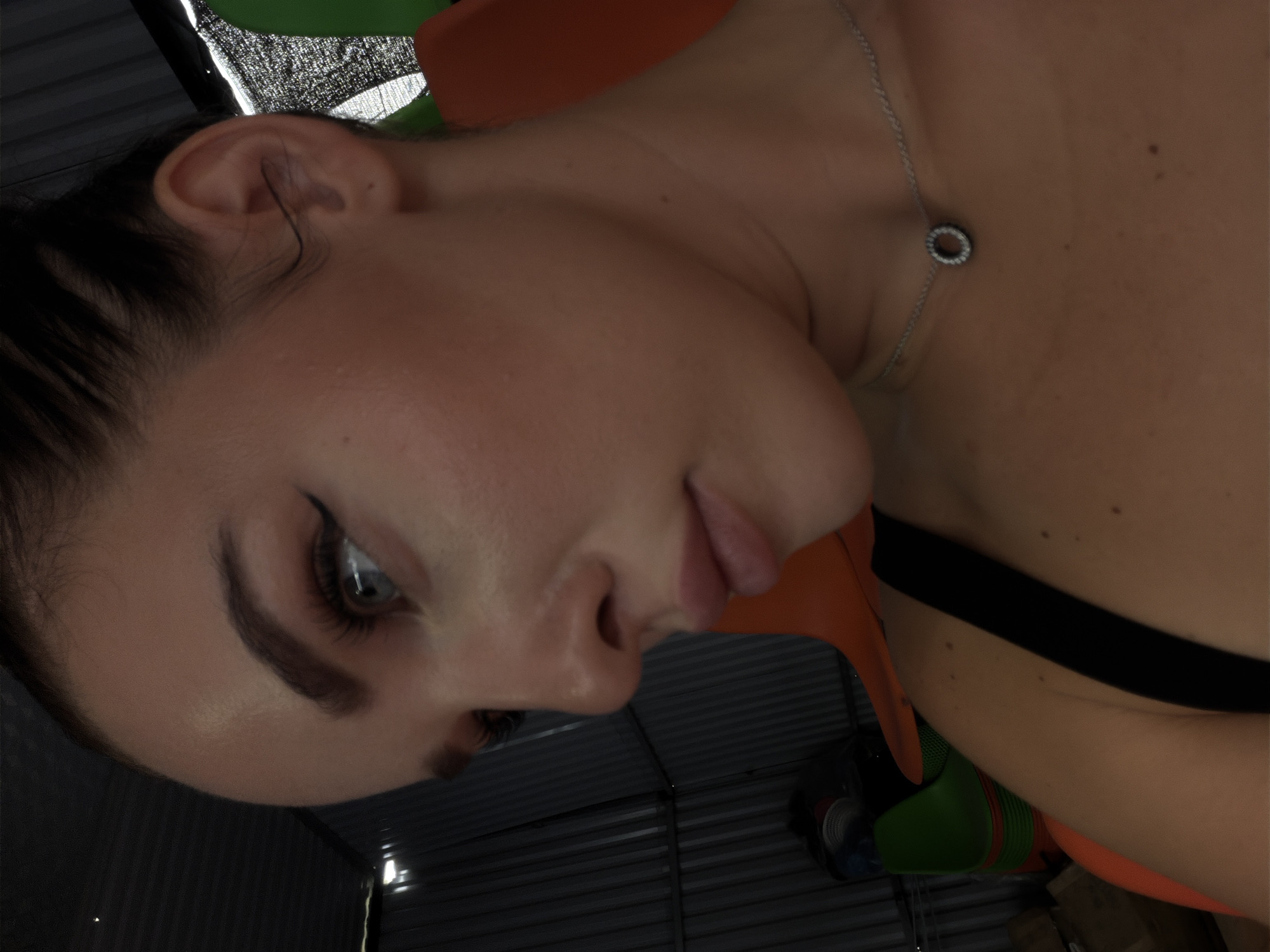 A close-up portrait features the left side of a person's face, heavily tilted to the right, against a dark and somewhat industrial background. The subject, appearing to be female, showcases clear skin, dark hair pulled back from her face, and a contemplative expression with her left eye gazing downwards. She wears winged eyeliner and has well-defined, dark eyebrows. Her lips are slightly parted. A thin silver chain necklace with a small, circular pendant adorns her neck, resting near her clavicle. A black strap, likely from a bra or top, is visible over her shoulder. The skin on her neck and shoulder shows a few small moles.
The background is predominantly dark, revealing textured surfaces, possibly corrugated metal or slatted panels, indicative of an indoor space such as a garage or workshop. Bright orange and green elements are also faintly visible in the background, suggesting furniture or equipment. The lighting is focused on the subject's face, creating soft highlights and shadows. No specific activity is depicted other than posing for the photograph, capturing an intimate and thoughtful moment. There is no visible text in the image. The scene is situated in Nam Nha Trang Ward, Vietnam.
