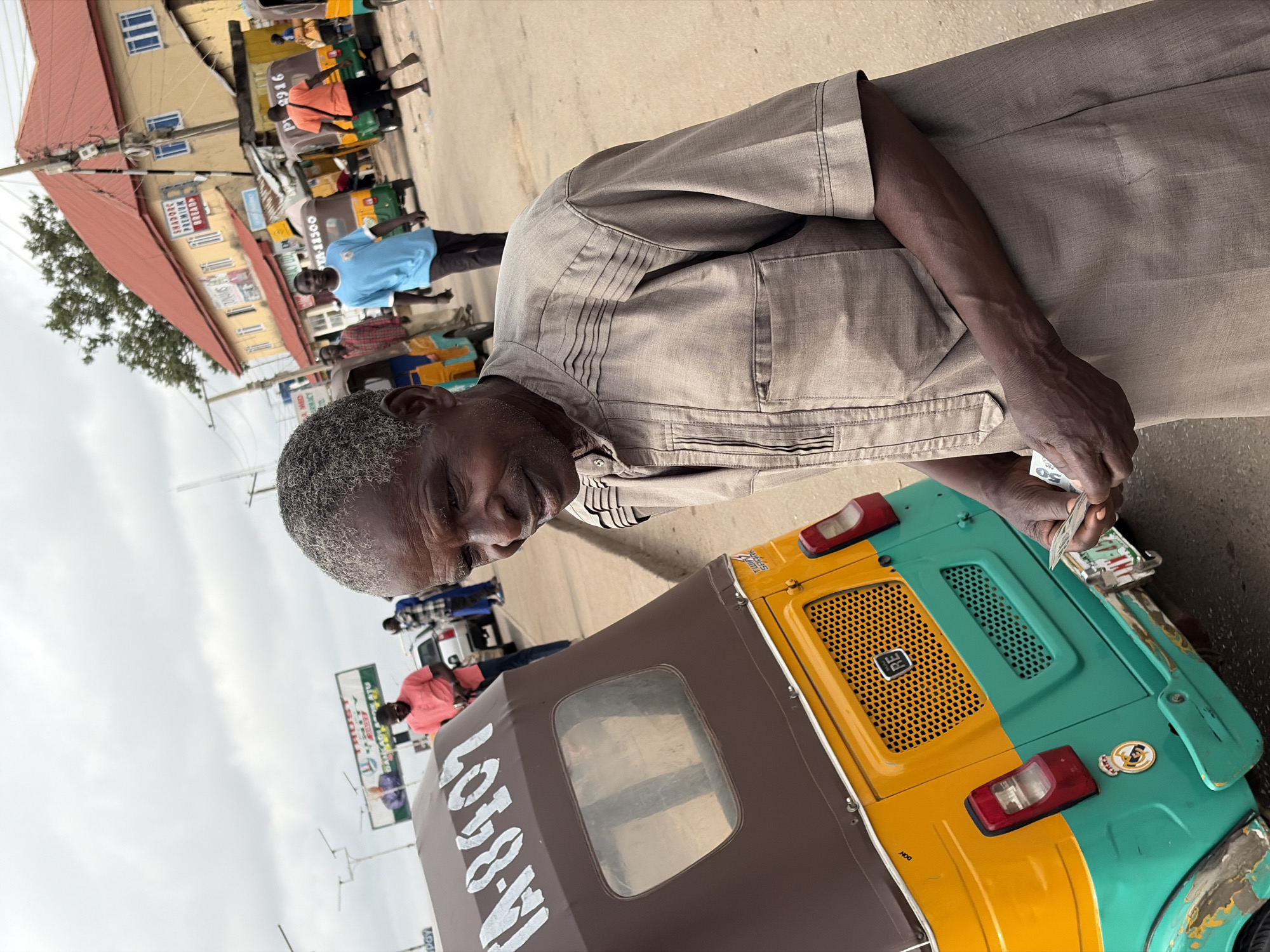 This media file captures a vibrant daytime street scene in Gura Top, Nigeria, under an overcast sky.
The central subject is an older man with dark skin and short, grey hair, wearing a light brown or tan long-sleeved traditional outfit with intricate stitching details on the chest and sleeves. He is positioned next to the rear of a three-wheeled auto-rickshaw, commonly known as a Keke Napep. The man is holding several Nigerian Naira banknotes, one clearly showing the denomination "50," suggesting a transaction is taking place, possibly payment for a ride or part of his work as a driver. His expression appears neutral to slightly thoughtful as he looks downwards.
The auto-rickshaw in the foreground is brightly colored, with its rear section split into yellow and green panels, and a brown canvas canopy over the passenger area. The grille on the yellow section has the letters "RE" visible, and "DON" is faintly seen below. A larger inscription, "LOTO-W," is visible on the brown canopy, alongside "TA-8407."
In the background, the street is bustling with activity. Other people, mostly men, are seen walking or standing. One man wears a light blue shirt, another an orange shirt. Several more three-wheeled vehicles are visible, some with numbers like "TA-11500" and "P19916" on their rear. The street is lined with buildings, some with distinctive red corrugated roofs, and several commercial signs are visible. Text on these signs includes "SHADORC PREMIUM," "TRUST," "TWI," "SAARK," and "MET." The overall scene depicts a typical busy thoroughfare in an urban or semi-urban setting in Nigeria.