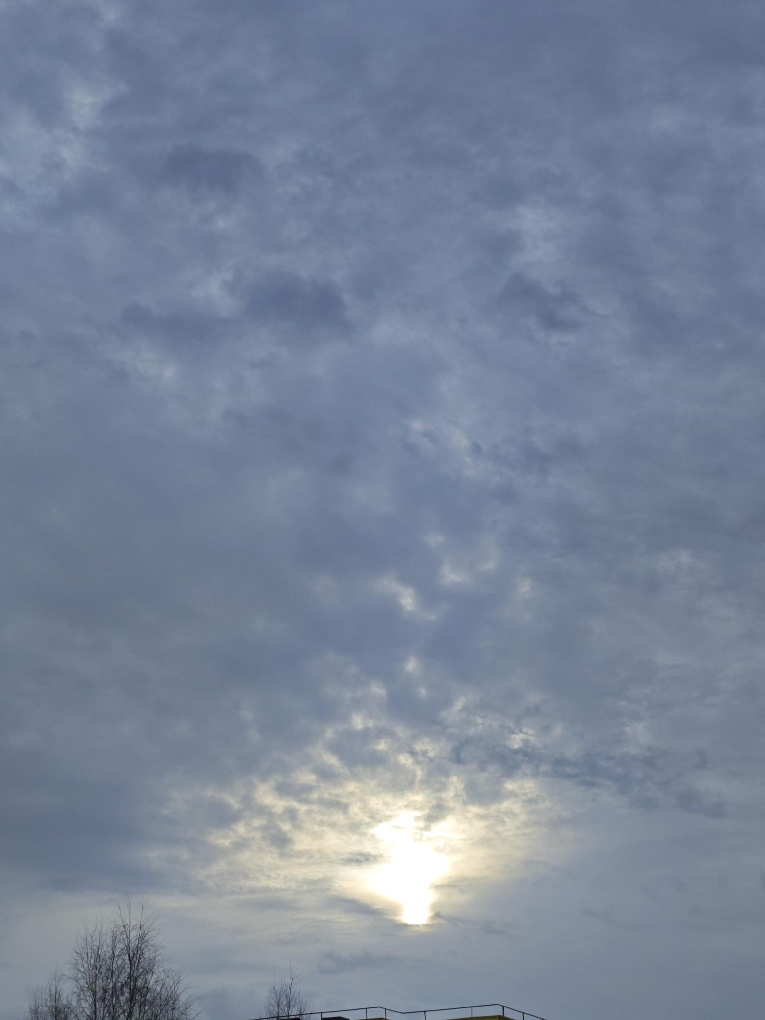 The image depicts a cloudy sky, likely in the Марковский area of Russia. The sun is obscured by the clouds, creating a bright spot in the lower center of the frame. In the bottom portion of the image, there is a partially visible structure or building and some bare trees.