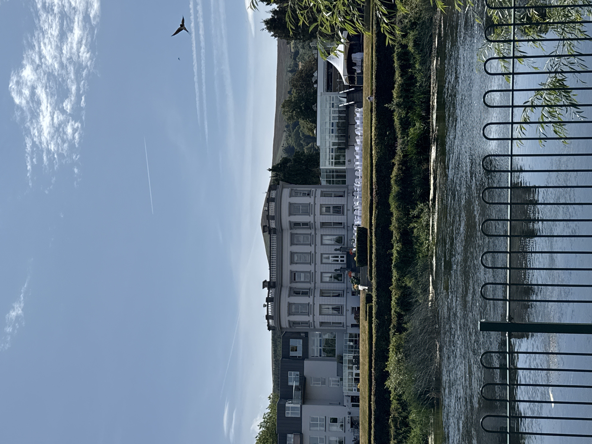This image depicts a view of the Clonmel Park Hotel in Clonmel, Ireland, from across a body of water. The hotel is a large, multi-story building with a grand, classical-style central section featuring a curved facade and numerous windows, attached to a more modern section with a flat roof and large glass windows. In front of the hotel, rows of white chairs are set up on a lawn, suggesting an outdoor event. A white marquee is also visible to the right of the hotel. The scene is framed by lush green hedges and trees in the foreground and middle ground. The river in front of the hotel reflects the blue sky, which has scattered white clouds and a contrail from an airplane. Two birds are visible in the sky. A dark metal fence runs along the bottom of the frame, separating the viewer from the river. The background features rolling green hills.