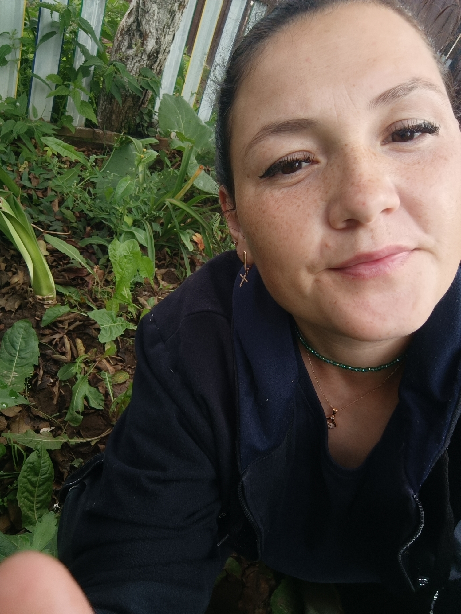 A woman is pictured in a close-up selfie outdoors, with greenery and foliage in the background. She has fair skin with freckles, dark hair pulled back, and is wearing makeup including eyeliner and mascara. She is wearing a dark navy jacket and a green beaded necklace with a gold pendant. The setting appears to be a natural environment with plants, leaves, and a tree trunk visible behind a light-colored fence. There is no indication of the city of Karsha, Russia in the image.