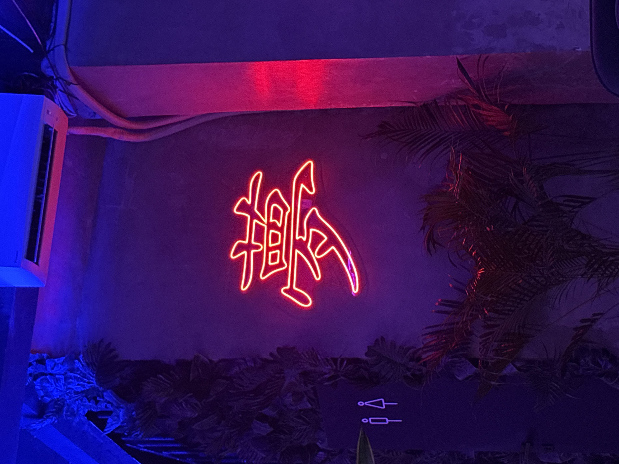 The image shows a dimly lit indoor setting, likely a bar or lounge, in Kuta Selatan, Indonesia. The color scheme is dominated by shades of blue, purple, and red. On the wall is a neon sign in a stylized Asian script. To the left is what appears to be part of an air conditioning unit, illuminated in blue. Below the neon sign, at the bottom of the image, is dark foliage or decorative greenery with toilet signs visible. The right side of the image features palm fronds. There are some cables and conduits visible near the top left.