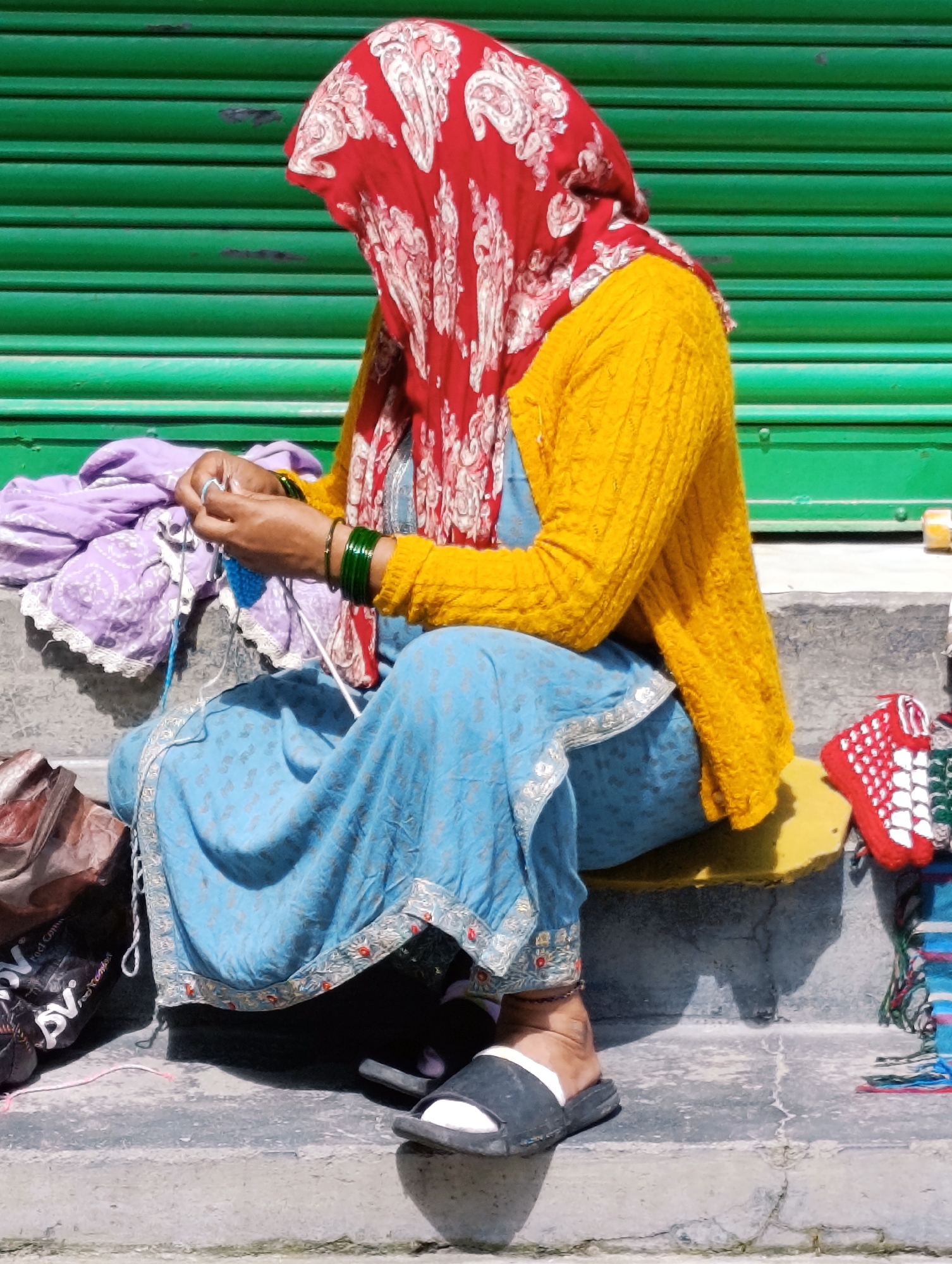 An individual wearing a red patterned headscarf, a yellow knitted cardigan, and a blue patterned skirt sits on concrete steps in Vashisht, India, knitting with blue and white yarn. To the left of the individual, a pile of purple fabric rests. To the right, a red knitted item with white accents and a colorful woven textile are visible on a yellow cushion. The background consists of a green corrugated metal surface.