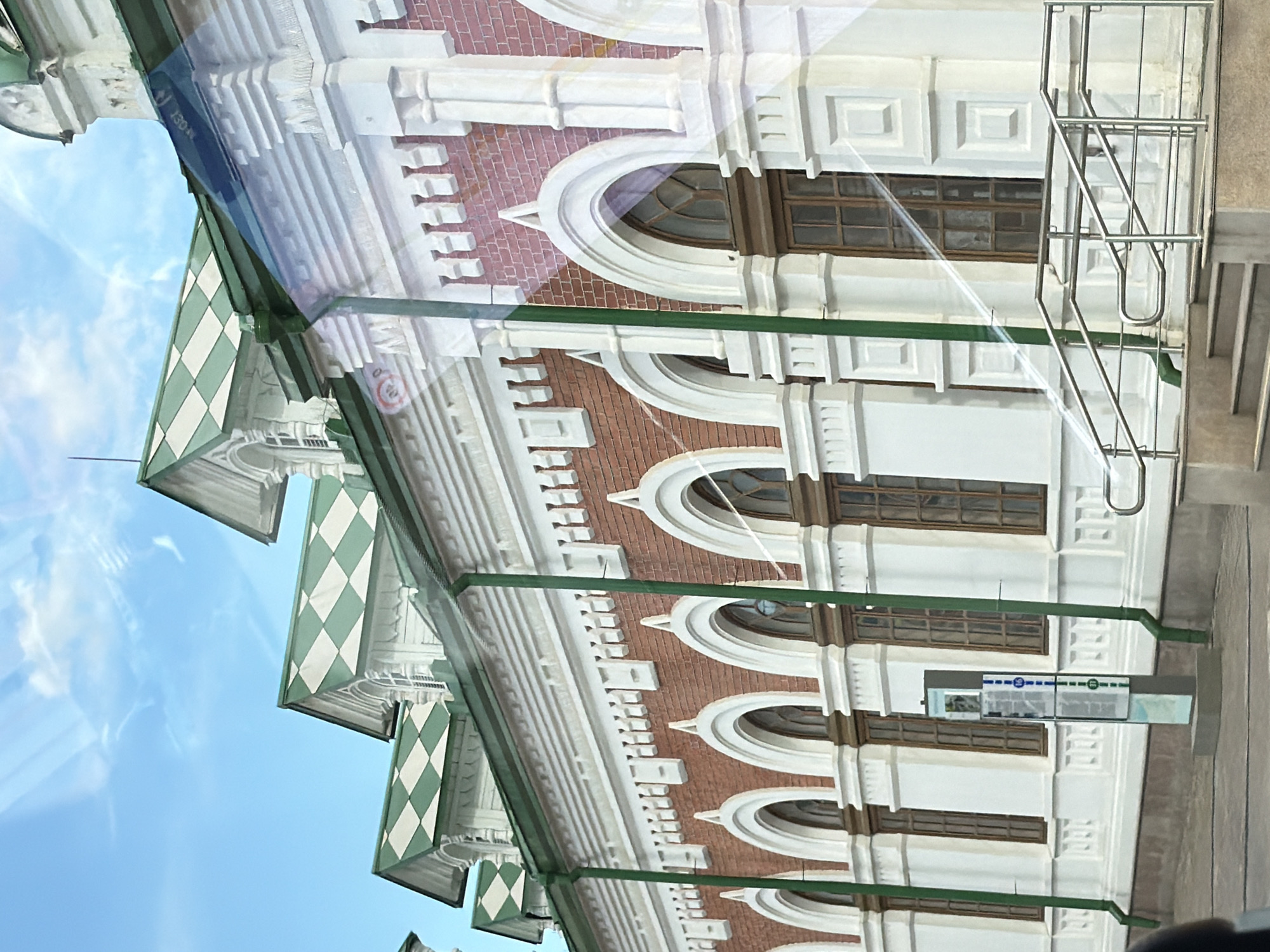 The image shows an exterior view of a building with classical architecture. The building has a white facade with decorative elements and red brick accents. Arched windows are visible, some with intricate wooden frames. The roofline features a green and white diamond pattern on inclined surfaces. A metal railing and staircase are attached to the building's exterior.
A sign with a blue and white design and some text is mounted on the building. Reflections on the building's surface suggest it might be made of glass or a reflective material, possibly indicating a modern addition to an older structure, or a window view of the building. The sky is visible in the background, with some clouds.
The overall impression is of an ornate and historic building, possibly a museum, government building, or a public institution. The lighting suggests it is daytime. There is no clear indication of specific activity or people, with the focus being on the architectural details and the building itself. The location context provided is Perm, Russia.