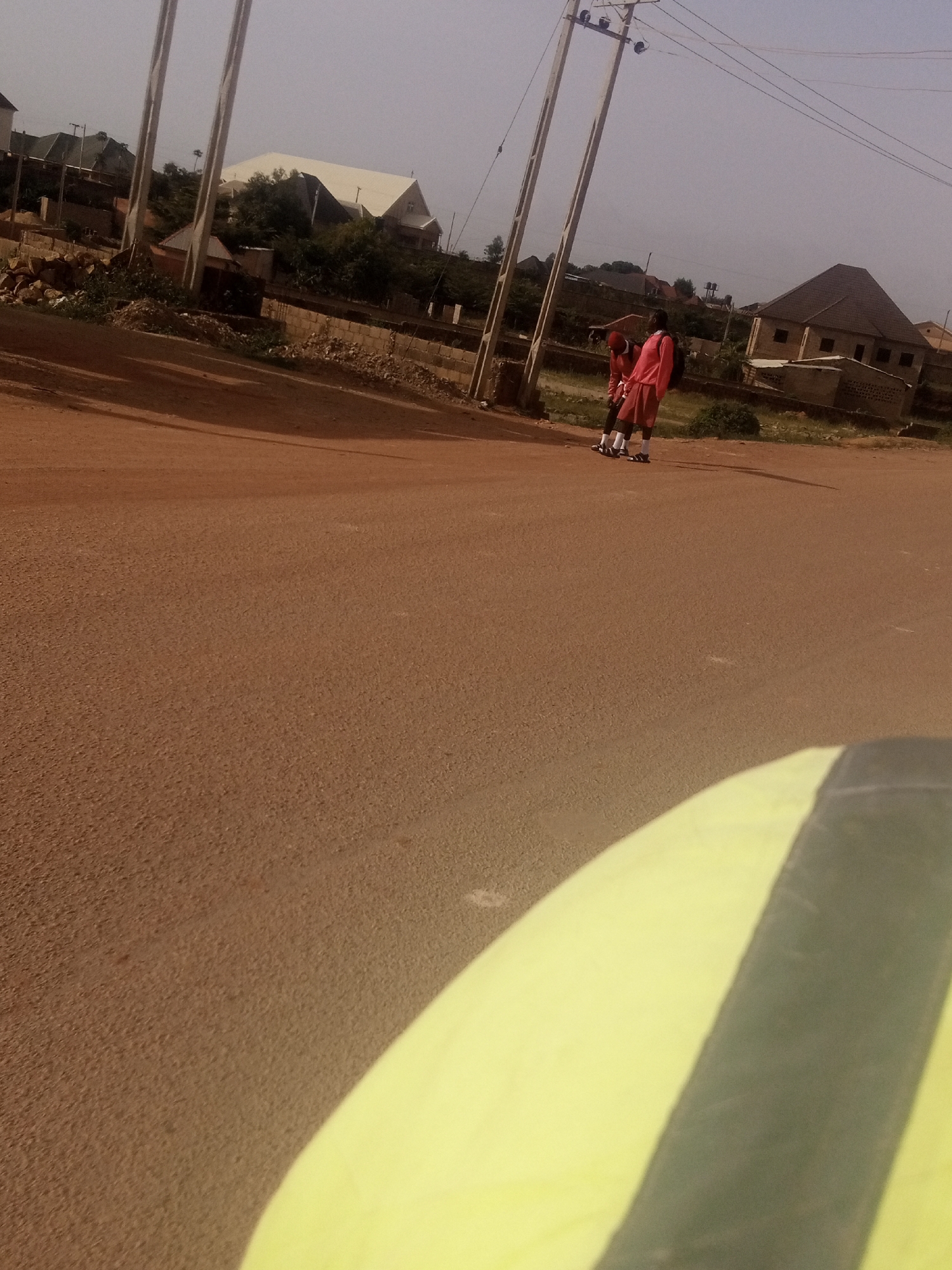 In Rayfield, Nigeria, two individuals, appearing to be of school age, are standing on a dirt road. One person is wearing a red top and shorts, while the other is wearing a red top and shorts. Both are wearing white socks and dark shoes. They are positioned near a utility pole with overhead wires. In the background, there are several buildings, including a structure with a prominent white roof that resembles a church. The ground is a reddish-brown dirt, and there are some scattered stones and vegetation. The scene appears to be a typical outdoor environment in a Nigerian town. A blurry, yellow object is in the foreground, partially obscuring the bottom of the image.