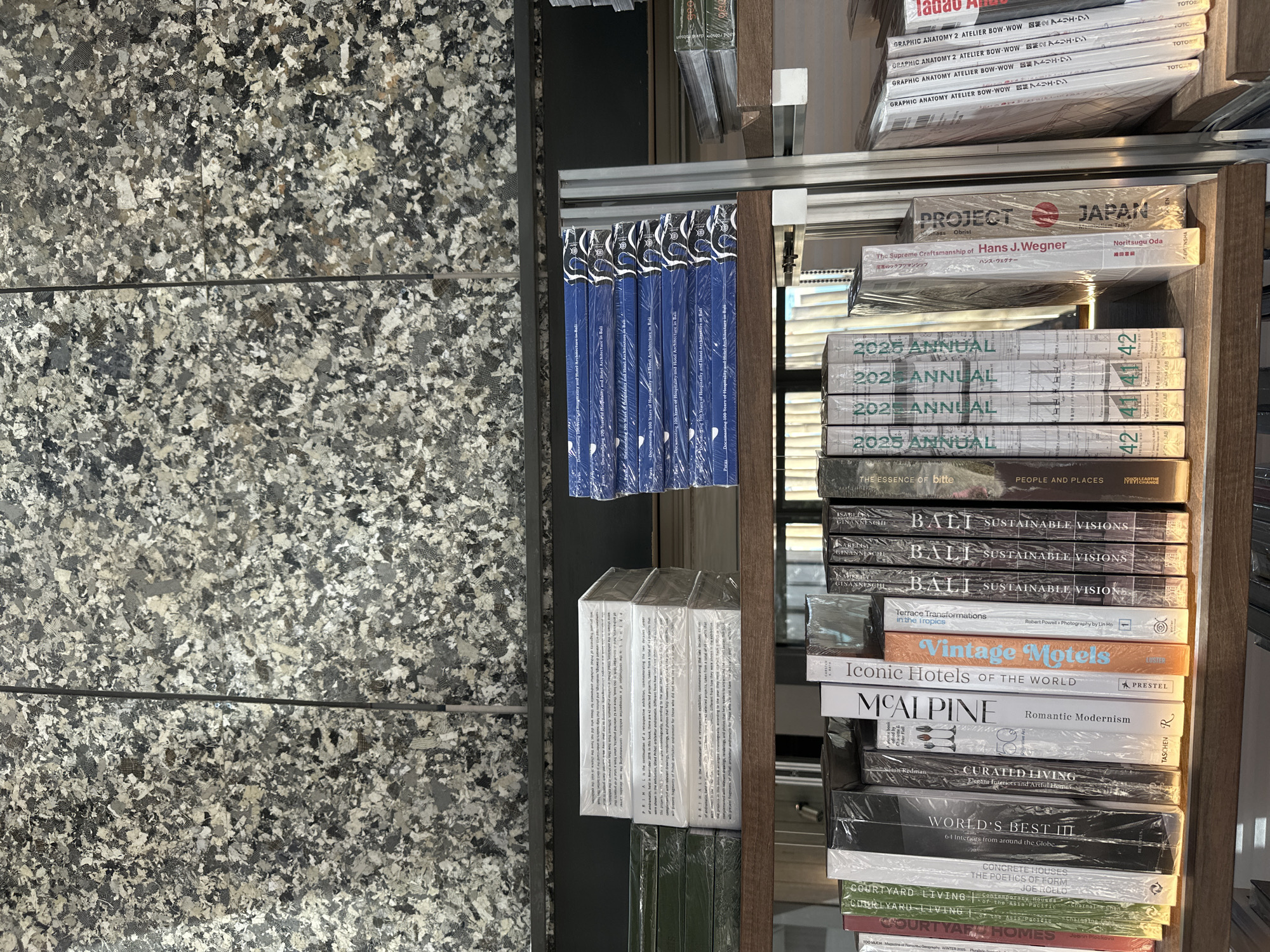 The image depicts a bookshelf filled with books, some stacked horizontally and others vertically. The background features a textured wall with a speckled pattern of gray, black, and white. The bookshelf itself is made of wood, with metal supports visible on the right side.
On the top shelf, horizontal stacks of white-covered books are present, some wrapped in plastic. Below them, green-covered books are also stacked horizontally and appear to be wrapped. To the right, a stack of blue books with white lettering is visible.
The bottom shelf displays a collection of vertically oriented books with various colorful covers and titles. Some of these books are also wrapped in plastic. Titles visible include "COURTYARD LIVING," "WORLD'S BEST IN," "MCALPINE," "Iconic Hotels of the World," "Vintage Motels," "TERRACE TRANSFORMATIONS," and several books with "BALI SUSTAINABLE VISIONS" and "2025 ANNUAL" on their spines. Other titles like "Project Japan" and "The Essence of Bliss" are also discernible.
There are no people or actions visible in the image. The environment suggests an indoor setting, possibly a bookstore, library, or gallery. The presence of books about Bali and architectural themes might indicate a thematic collection. The reference to Denpasar, Indonesia, suggests the location of this establishment.