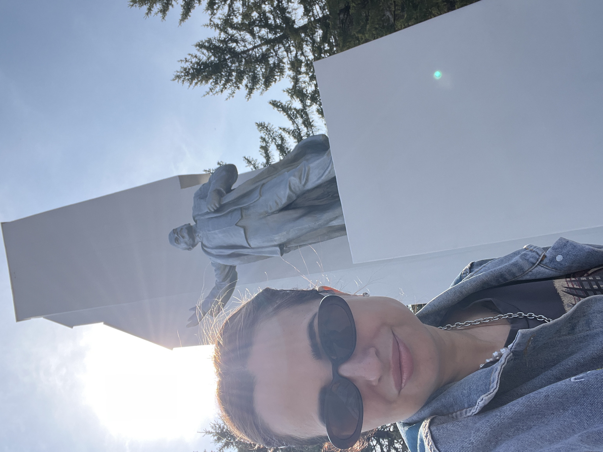 A woman wearing a denim jacket and sunglasses is taking a selfie in front of a monument featuring a statue of a man in a long coat. The statue is mounted on a multi-tiered white structure. The background includes trees and a bright sky with the sun shining. The location is in the city of Unknown, Unknown.