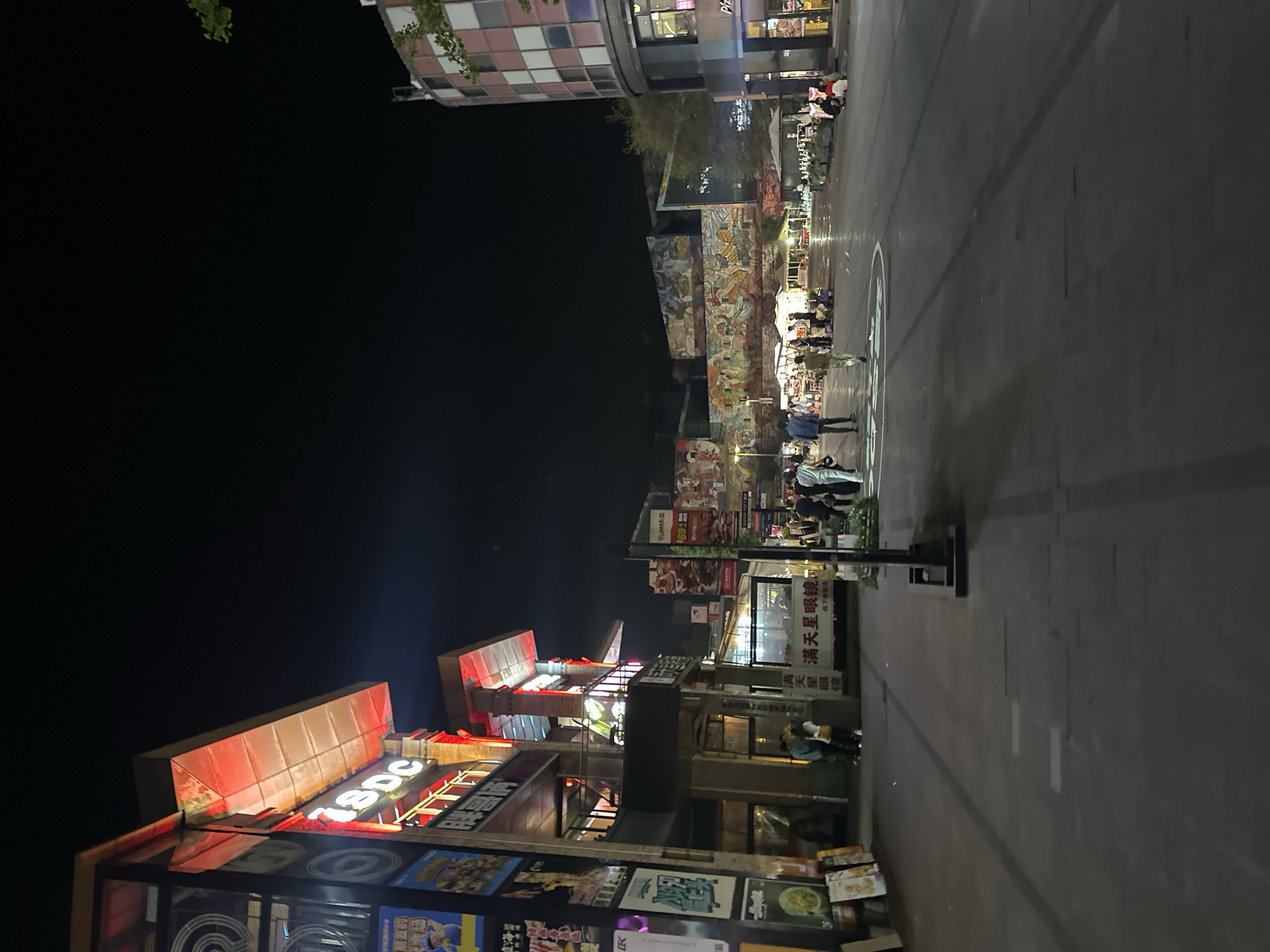 The image shows a street scene at night in Chongqing High-tech Zone, China. Several people are walking on the sidewalk. There are several commercial establishments on both sides of the street with neon lights. On the left is a building with a red sign. There is another building decorated with various painted and illuminated posters. There is a street light post with a sign that reads, "Mantianxing Glasses." Further down the street are a few stalls and more illuminated structures and figures of people. There is a gray-colored paved road occupying the right half of the image. In the far distance is a building with square tile designs. The sky is predominantly black.