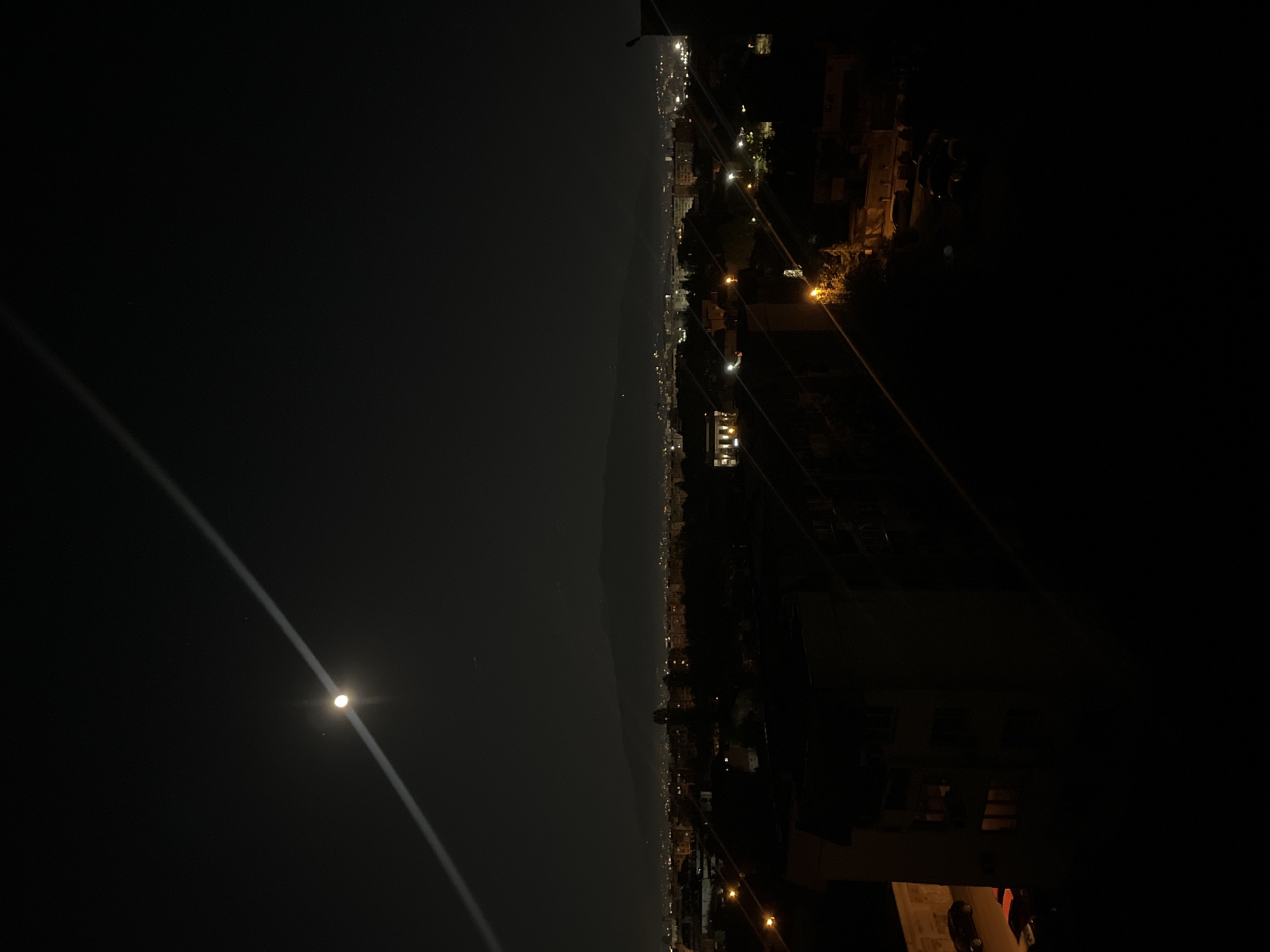 The image depicts a nighttime urban scene, likely from a high vantage point. The right side reveals a city with illuminated buildings and streetlights, suggesting urban development. A coastline or large body of water is visible on the left, illuminated by a bright moon. No people are discernible. The location is potentially Sofia, Bulgaria, based on the urban landscape and coastal proximity, but this is speculative and requires further verification.