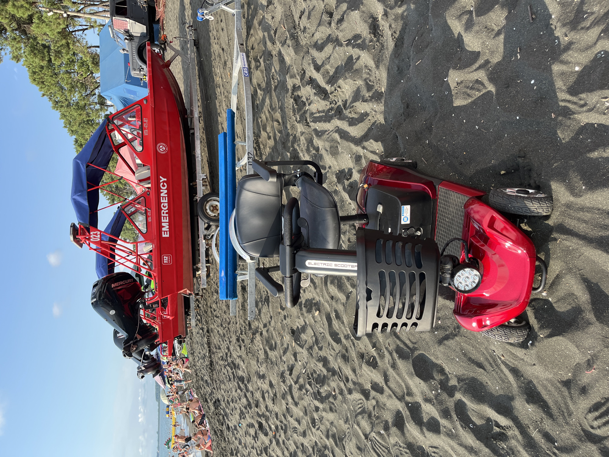 In Ureki, Georgia, on a dark sand beach, a red mobility scooter is parked in the foreground. Behind it, a red emergency boat with two outboard motors is situated on a trailer. The boat has a blue canopy and the word "EMERGENCY" is visible on its side. Further in the background, people are visible on the beach and in the water, and trees line the horizon. The sky is clear and blue, indicating a sunny day.