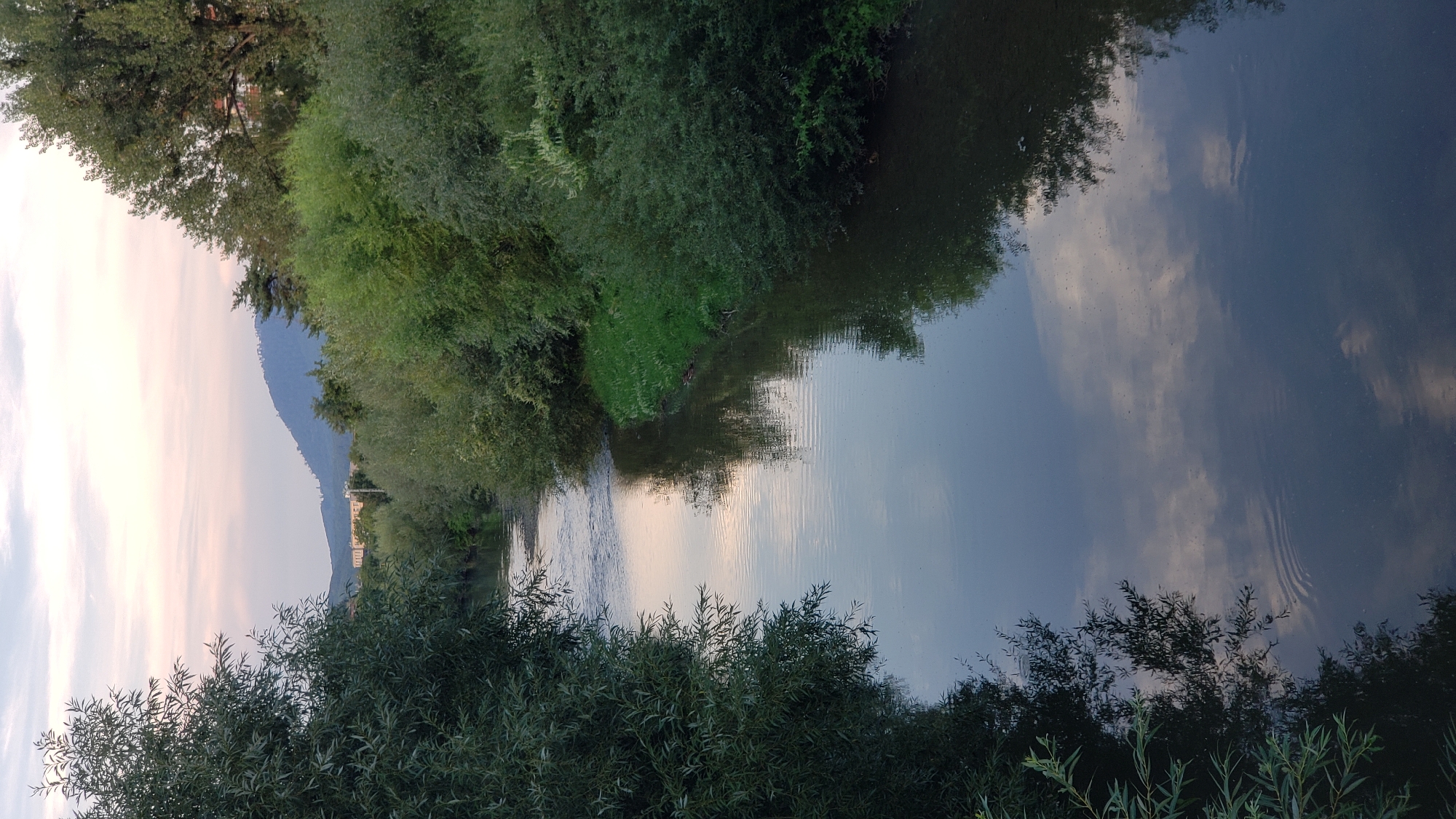This image depicts a serene river flowing through a natural setting, possibly on the outskirts of Prešov, Slovakia. The river is calm, with gentle ripples disturbing its surface, and reflects the soft hues of the sky. Lush green foliage lines both banks of the river, with trees and bushes creating a natural canopy. In the distance, a gentle mountain range can be seen under a twilight sky painted with pastel shades of pink and blue, with scattered clouds. A building complex is visible on the lower slopes of the mountains, suggesting proximity to an urban area. The overall atmosphere is tranquil, capturing a moment of natural beauty.