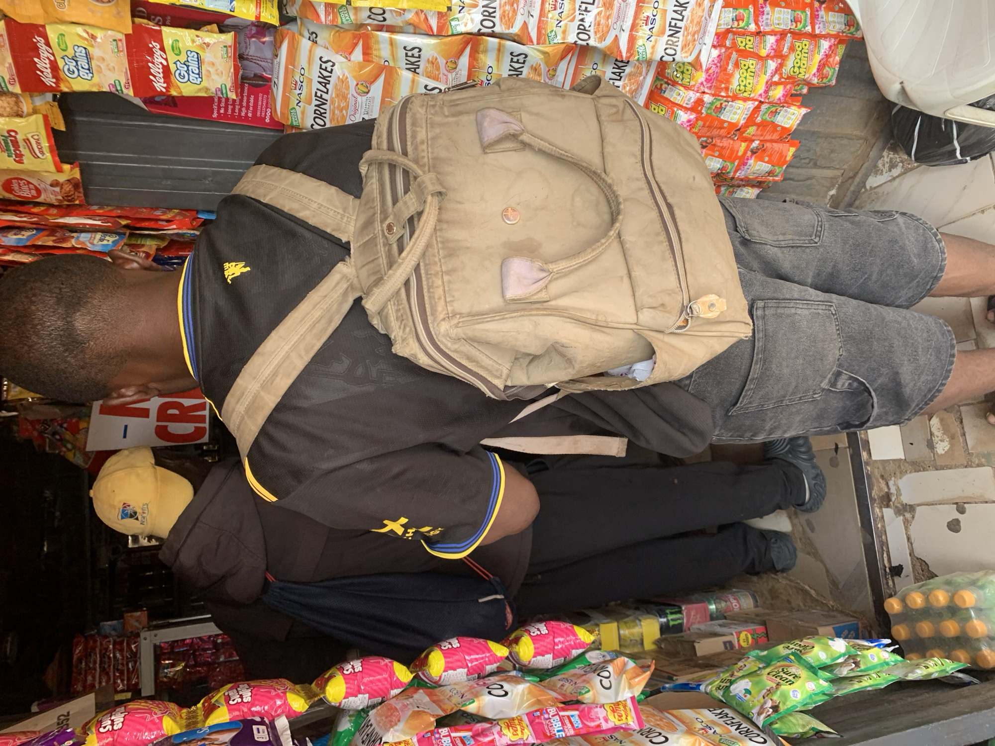 A man with a backpack stands in front of a kiosk laden with packaged food items in Rafin Par, Nigeria. He is wearing a black shirt with gold and blue accents, gray cargo shorts, and black shoes. The backpack is light brown with multiple compartments and straps. The kiosk shelves are stocked with various brands of cornflakes, snacks, and drinks, including Kellogg's and Nasco products.