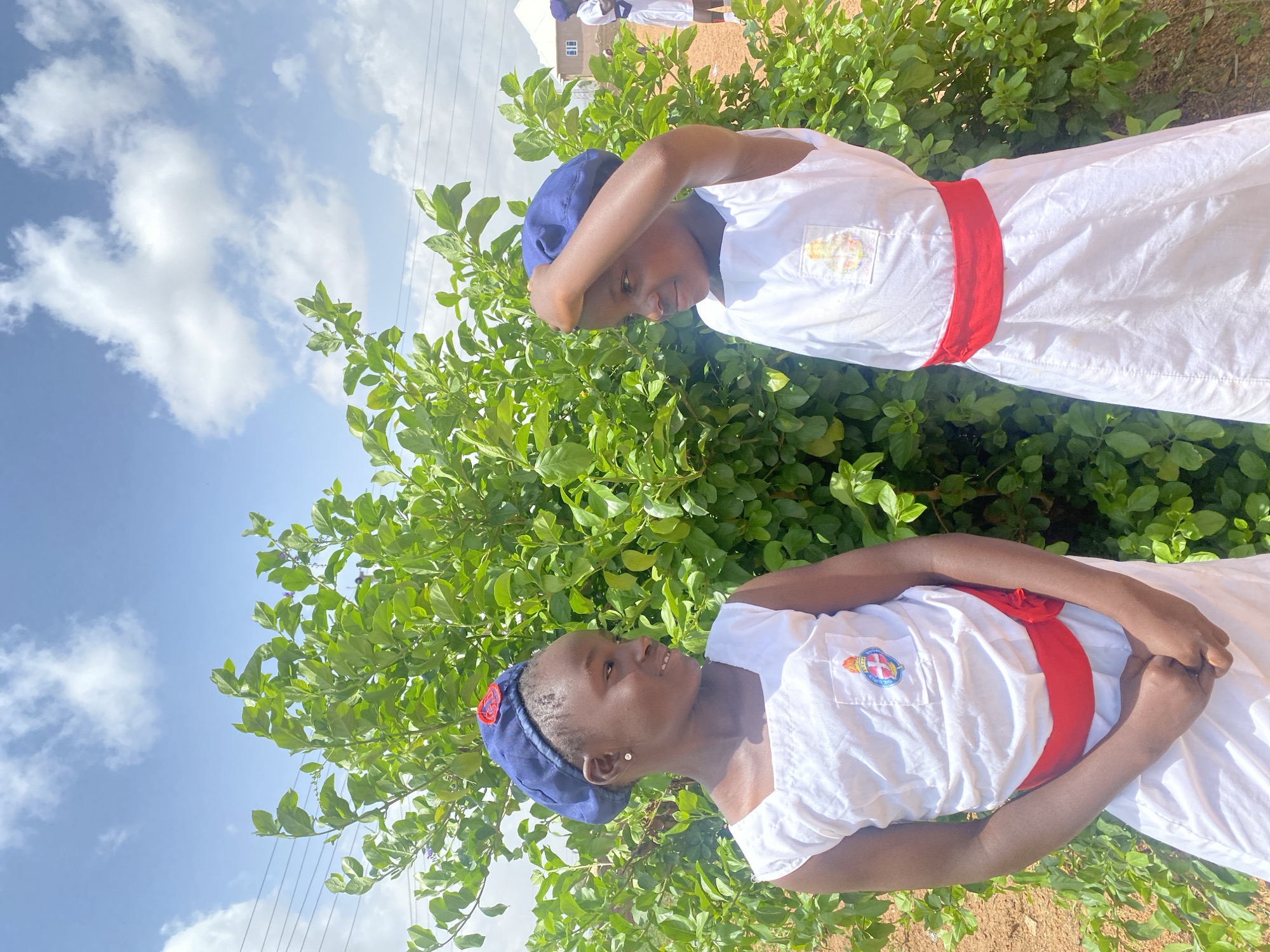 Two young people are dressed in white uniforms with red sashes, standing outside amidst green shrubs under a partly cloudy sky. Both are wearing navy blue caps. The location appears to be in Turu, Nigeria, though specific city details are not distinctly visible.