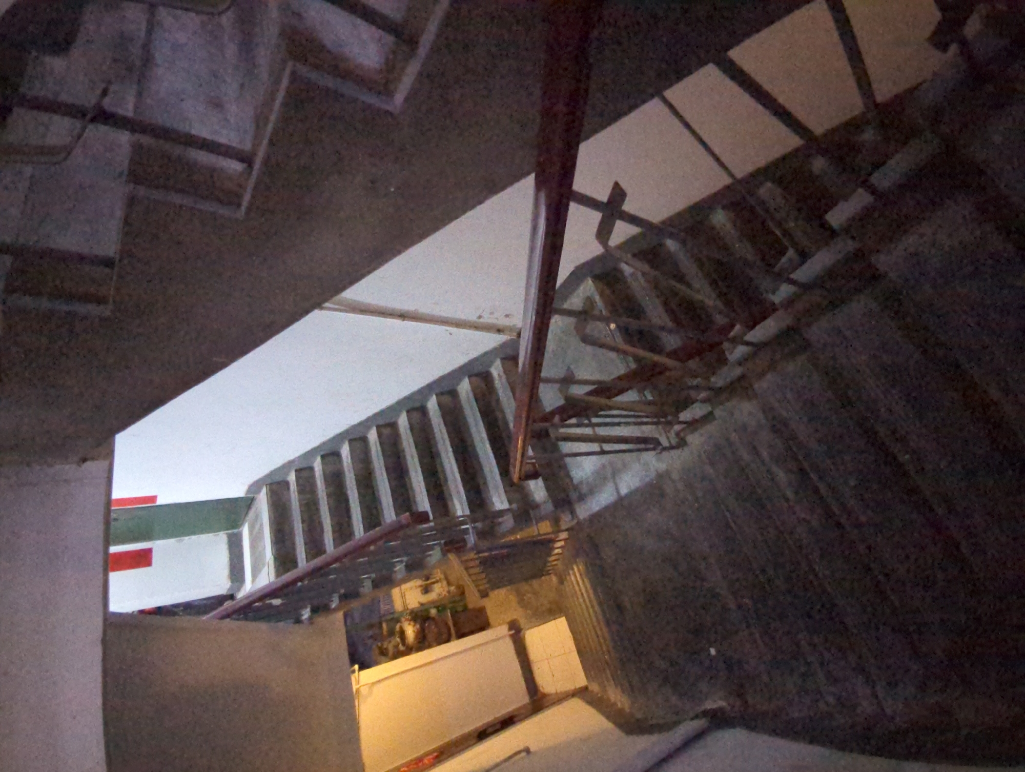 An indoor staircase with concrete steps and dark metal railings is visible from a high angle. The staircase is dimly lit, with some areas illuminated by artificial light. In the upper portion of the image, there is a glimpse of a doorway with red and green accents. The lower portion shows the continuation of the staircase and a section of a hallway with a light-colored wall. The environment appears to be a residential or commercial building. There is no indication of the location being in Yuzhong District, China.