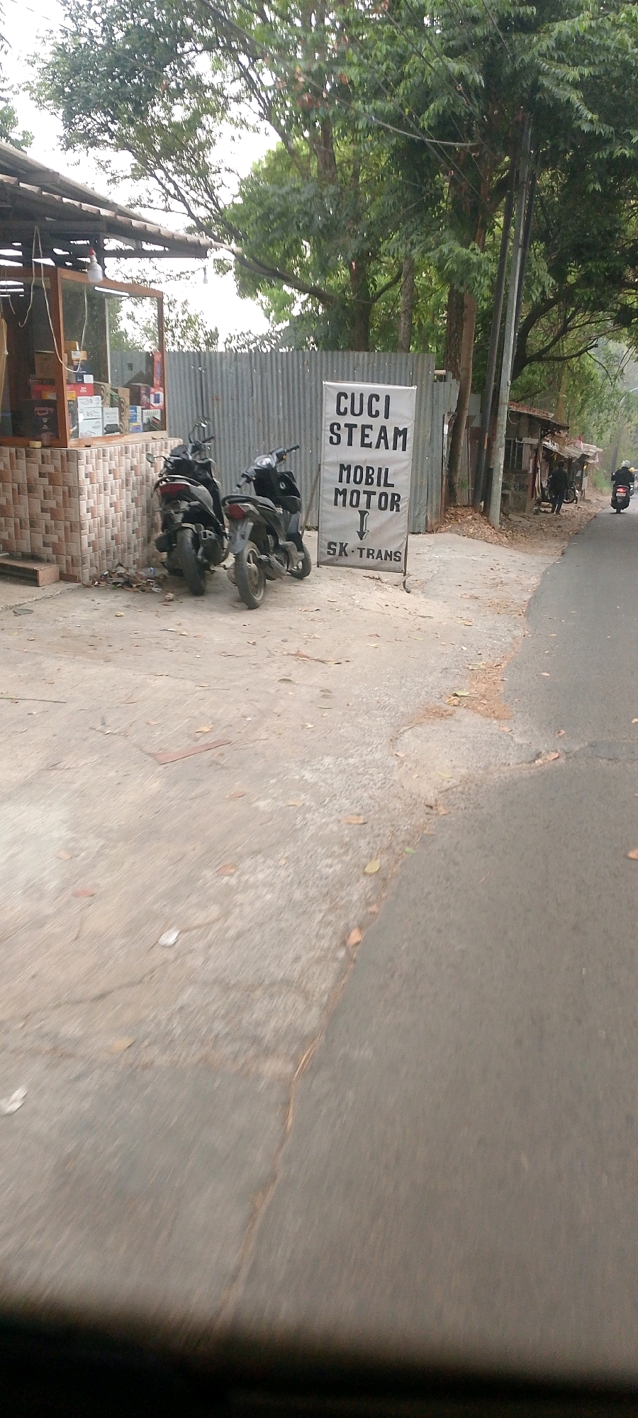 The image shows a street scene in what appears to be an urban or semi-urban area, likely in Bandung City, Indonesia, given the text on the sign. In the foreground, a cracked concrete surface with scattered leaves runs alongside an asphalt road. To the left, there's a small building with a corrugated metal roof, possibly a shop or stall, with shelves stocked with various items. In front of this structure, two parked motorbikes are visible. A large vertical sign stands near the motorbikes, displaying text in Indonesian: "CUCI STEAM MOBIL MOTOR" with an arrow pointing down to "SK TRANS". Beyond the building and sign, trees with dense foliage form a green backdrop. Further down the road, a small dilapidated structure with a thatched or corrugated roof is visible. A person on a motorbike is seen in the distance on the right side of the road, heading away from the camera. The overall scene suggests a commercial area situated alongside a road, possibly a car and motorbike wash service.