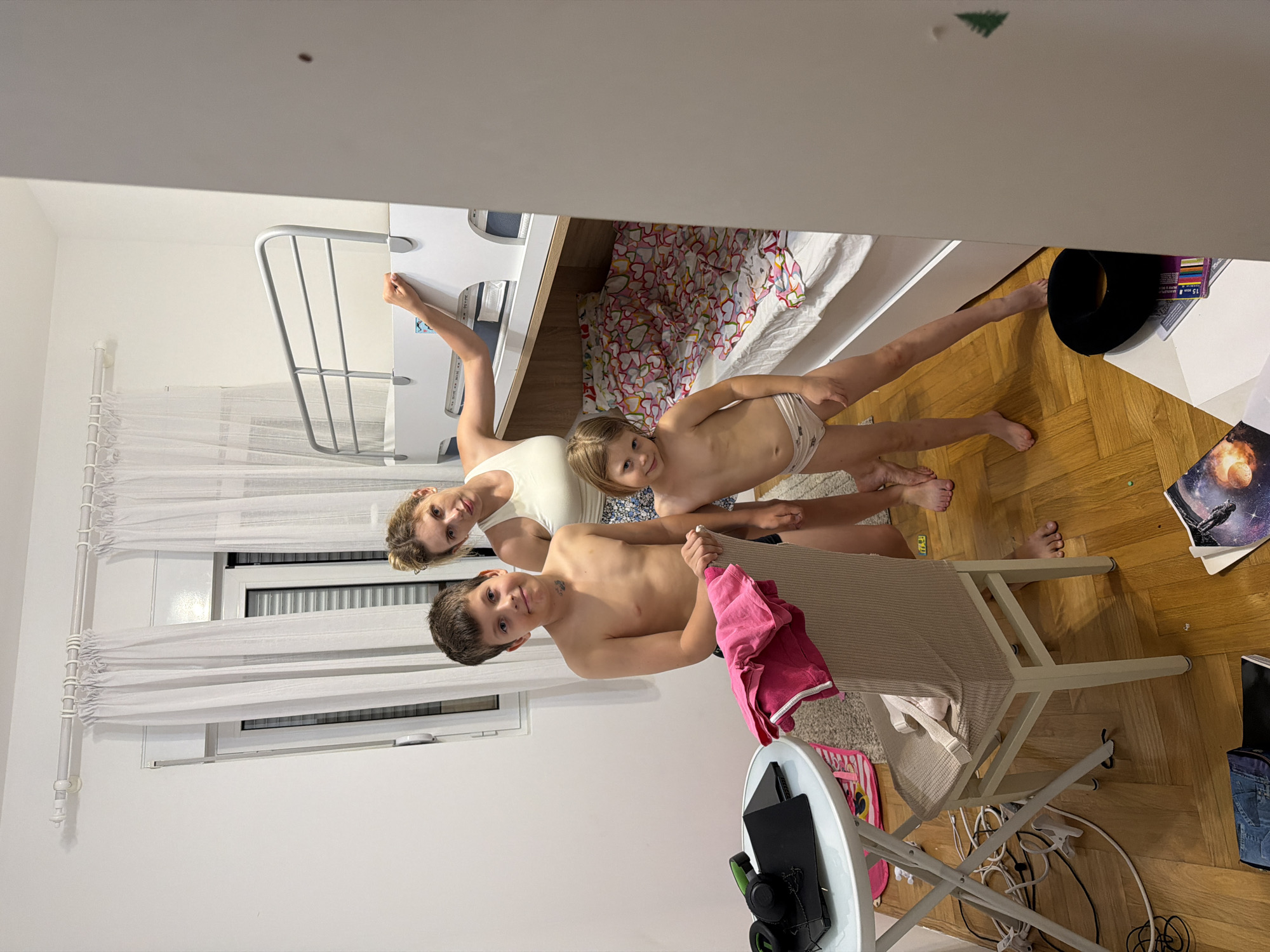 The image shows three shirtless people, two children and one adult female, standing in what appears to be a bedroom. The adult is on the left, with the two children to her right. The children are positioned in front of her, and they are all looking at the camera. The children are a boy and a younger girl. The scene includes a bunk bed with a colorful duvet, white curtains, and a wooden floor. There's a chair in the foreground, and various items are scattered around the room, including what looks like a book or magazine with a space-themed cover, a black hat, and some electronics. The overall impression is casual and domestic. The lighting suggests it could be daytime, possibly late afternoon or evening, as there is artificial light from a lamp or overhead fixture visible. The environment appears to be a home in Budva, Montenegro. The people have neutral expressions.