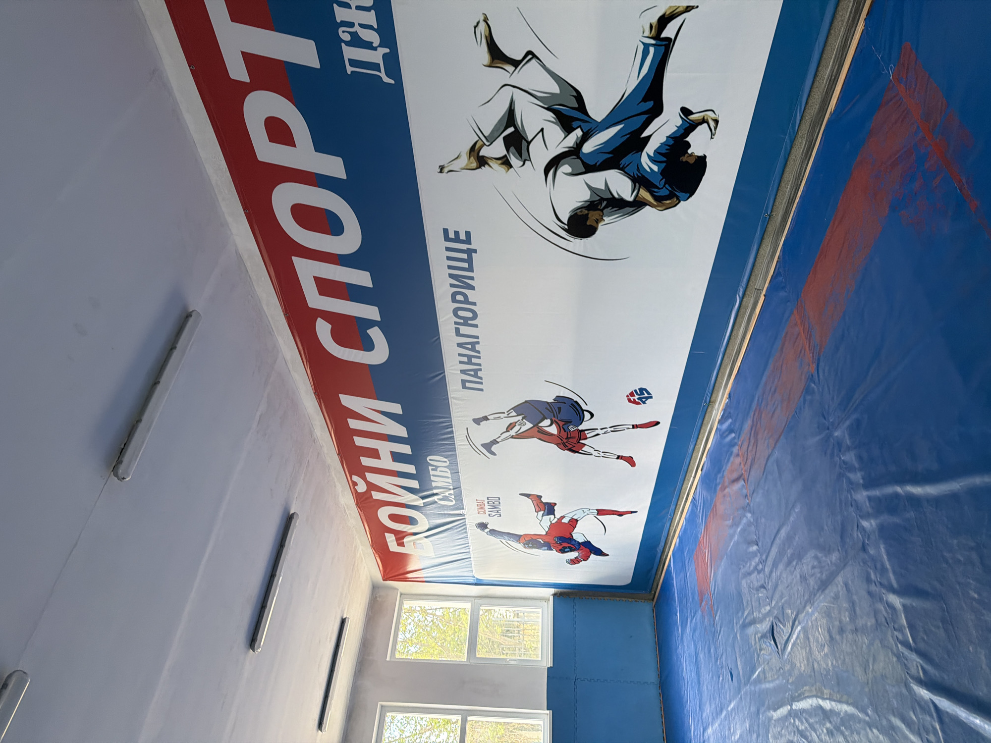 An indoor gym is shown from an eye-level perspective. The gym is located in Panagyurishte, Bulgaria.
The walls are painted light gray. There are light fixtures installed on the walls, in the form of fluorescent tubes. A window is located on the left side of the image, through which a green tree can be seen.
A large banner is mounted to the ceiling. The banner is decorated with images of martial artists in action. The words "Бойни Спорт" are written vertically in large, white letters with red shadow. Next to this text are more illustrations, specifically of wrestling combat. The words "ПАAHАЮРИЩЕ" are written below the graphics.
The floor is covered with blue mats with red details.