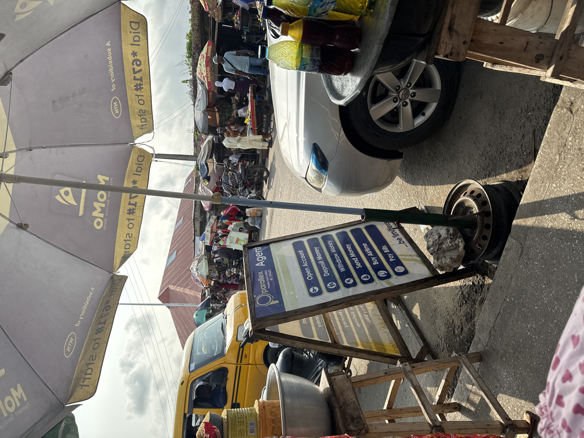 This is a daytime, street-level, medium shot of a market scene in Badagry, Nigeria. The foreground is dominated by an A-frame sign for "Parallax Agent," listing services such as "Open Account," "Deposit Money," "Withdraw Money," "Send Money," "Buy Airtime," and "Pay Bills." To the left of the sign, a large silver bowl sits on a wooden stand, next to a yellow van. In the mid-ground, a silver sedan is parked. The background reveals a busy marketplace with numerous stalls and people. Several umbrellas are open, providing shade over the vendors and shoppers. The overall environment suggests a vibrant commercial area.