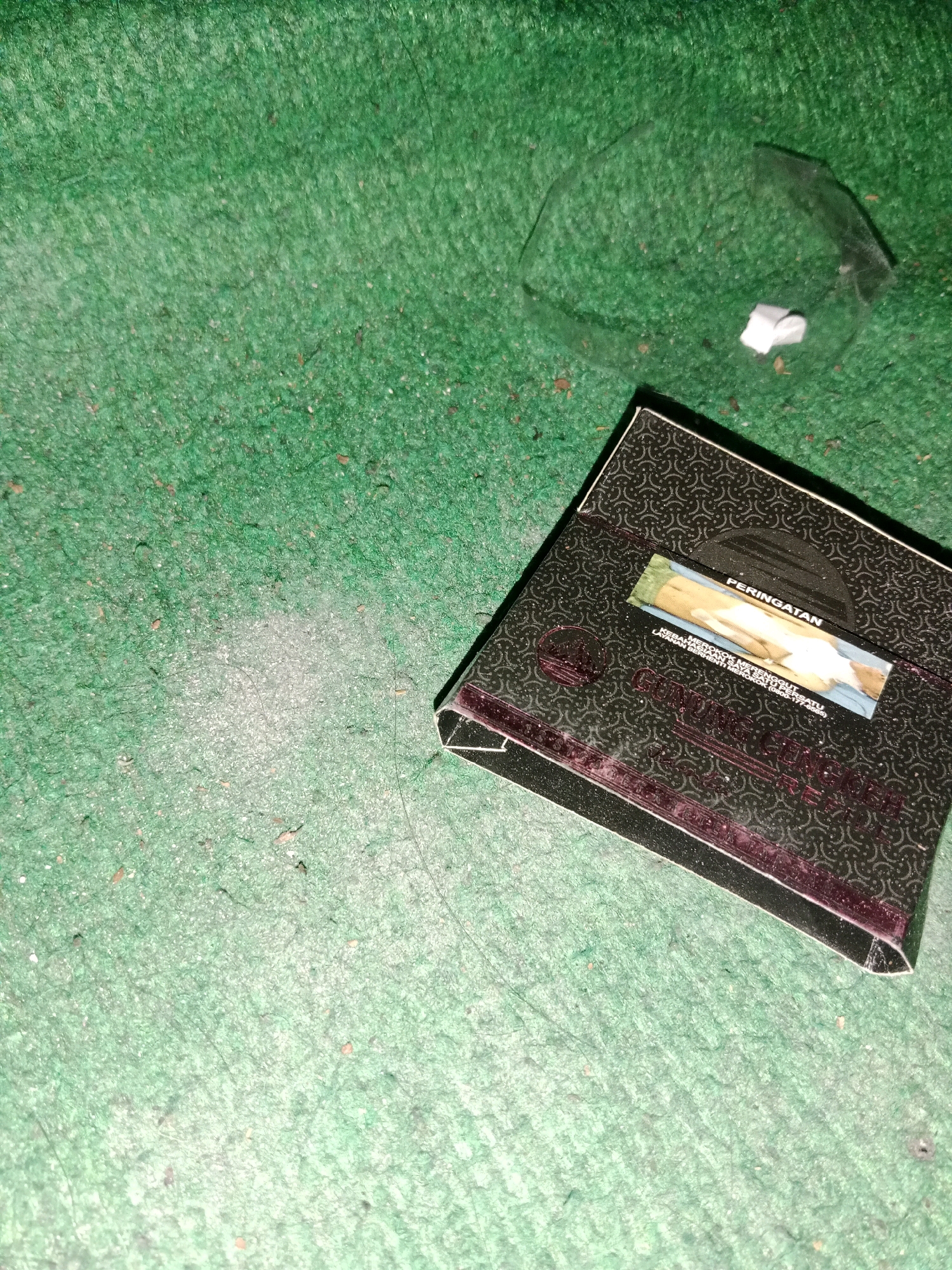 The image depicts a close-up, low-angle shot of a black cigarette pack with a warning label that reads "PERINGATAN" in white text. The pack is positioned on a textured green surface, likely a carpet or mat, which appears worn and contains scattered debris. To the upper right of the cigarette pack, a piece of clear, broken plastic is visible, along with a small white object. The lighting in the image is dim, casting shadows and creating a somewhat grainy texture. The context of Ringinrejo, Indonesia, is not visually represented in the image itself.