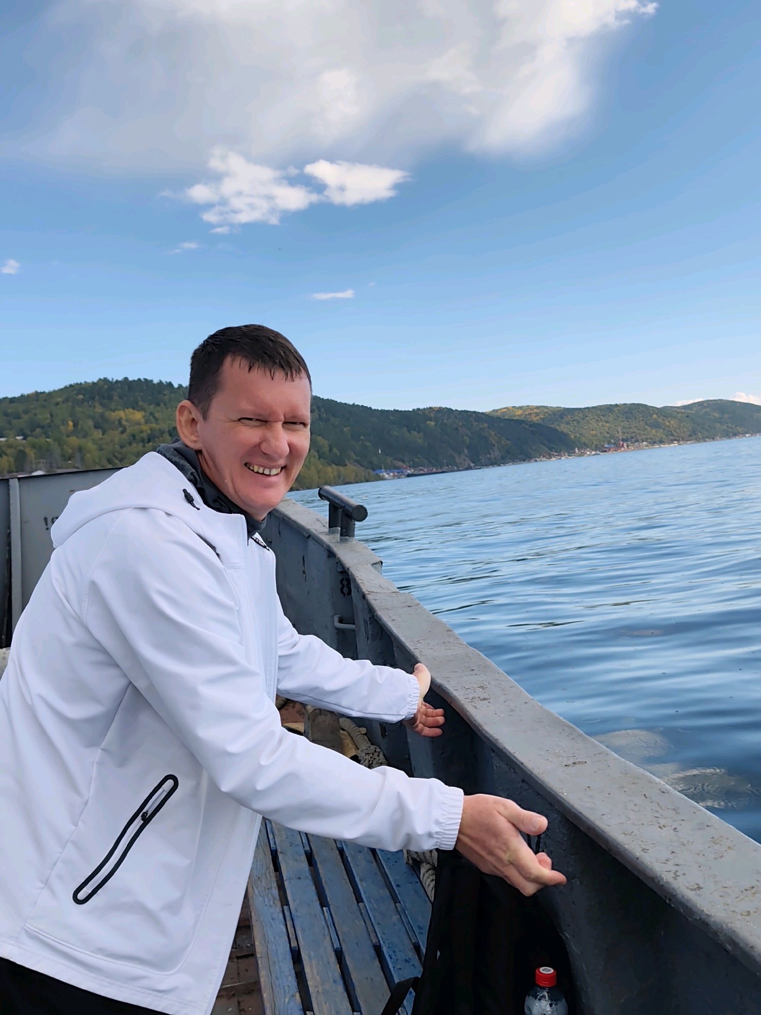 A man in a white hooded jacket is on a boat, smiling at the camera. He is positioned on the left side of the frame, leaning towards the right. The boat's interior shows blue wooden benches and a metal railing. The man's hands are extended outwards, touching the railing. Behind him, the water of Lake Baikal is visible, with a distant shoreline featuring green and yellow trees and a few buildings. The sky is blue with scattered white clouds. The scene suggests a day trip on the lake in Listvyanka, Russia, likely during the autumn due to the foliage colors.