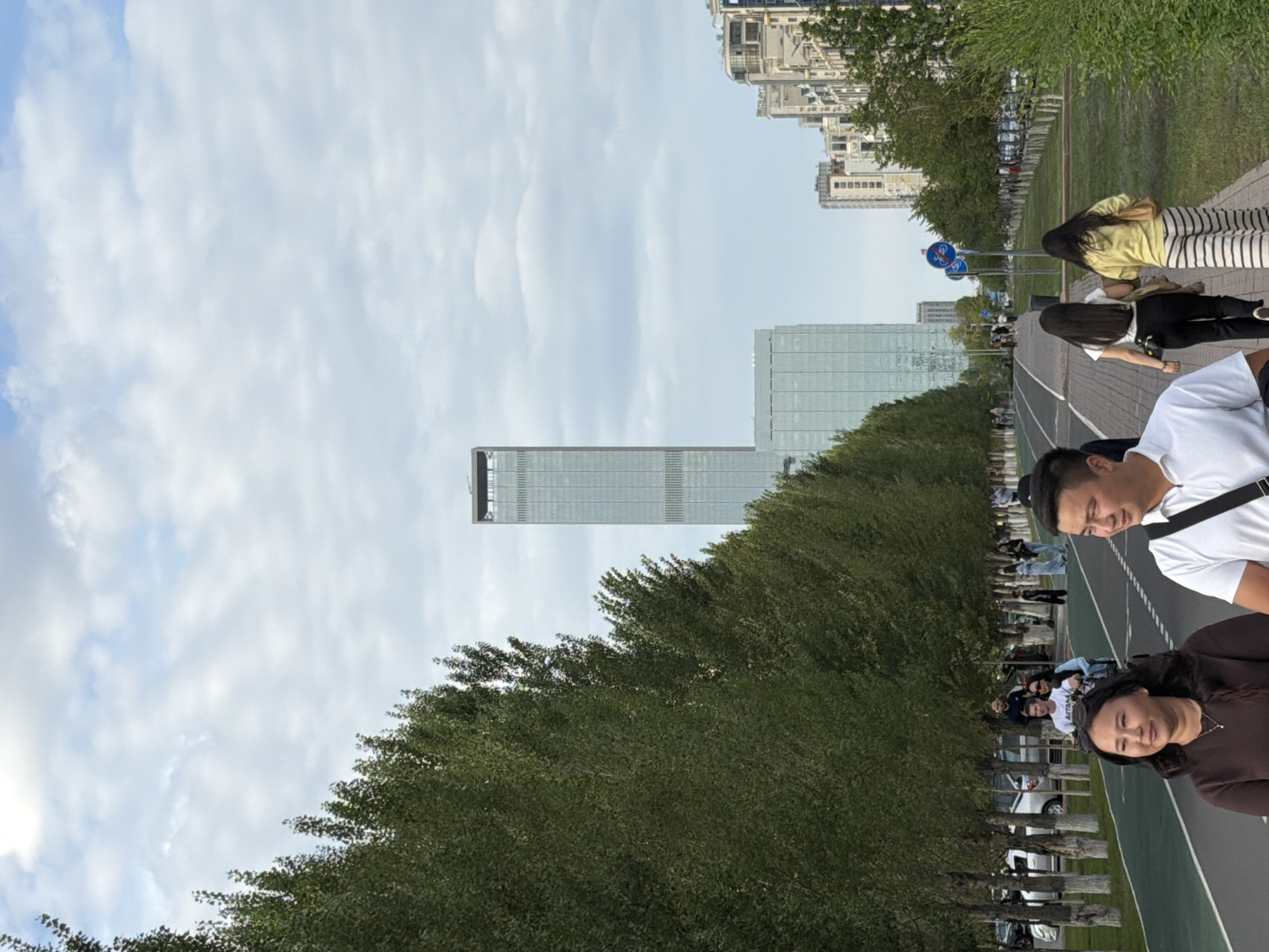 In Astana, Kazakhstan, a wide pedestrian walkway runs alongside a tree-lined avenue. A tall, modern skyscraper dominates the background, with other urban buildings visible further down the street. Several people are walking along the path, including a couple in the foreground, two women in the midground walking away from the camera, and various individuals scattered throughout the scene. A bicycle lane is present to the left of the walkway, and cars are parked along the side of the avenue. The sky is partly cloudy, suggesting it is daytime.
