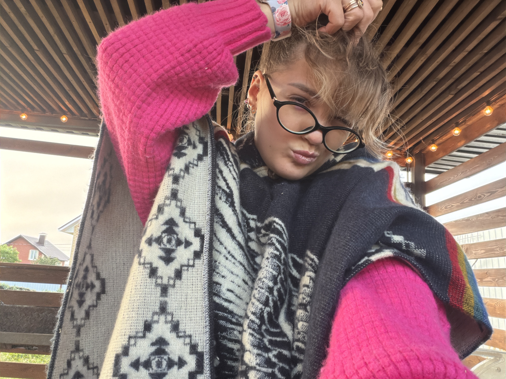 A medium-shot, slightly low-angle selfie captures a person, likely female, under a wooden slatted roof structure, possibly a gazebo or pergola. The person is looking directly at the camera with a playful, pouty expression, wearing black-rimmed glasses, and has one hand raking through their blonde or light brown hair.
They are dressed in a vibrant, textured hot pink sweater, which is visible on both arms. Over the sweater, they wear a patterned poncho or shawl, featuring intricate geometric designs in black, white, and grey, resembling tribal or Aztec patterns. A small portion of the shawl also shows stripes of color including blue, red, and yellow. On their left wrist, a watch with a floral pattern is visible, and on their right hand, several rings are worn on different fingers.
The background shows a bright, somewhat overcast sky. To the left, distant residential houses with reddish-brown roofs and light-colored walls are visible, suggesting an outdoor, suburban or rural setting. A wooden railing is also partially visible in the lower left. Above the person, the wooden slats of the roof run diagonally across the frame, with several string lights, emitting a warm glow, hanging underneath.
The image conveys a casual, personal moment, possibly captured during pleasant weather in Большая Шильна, Russia.