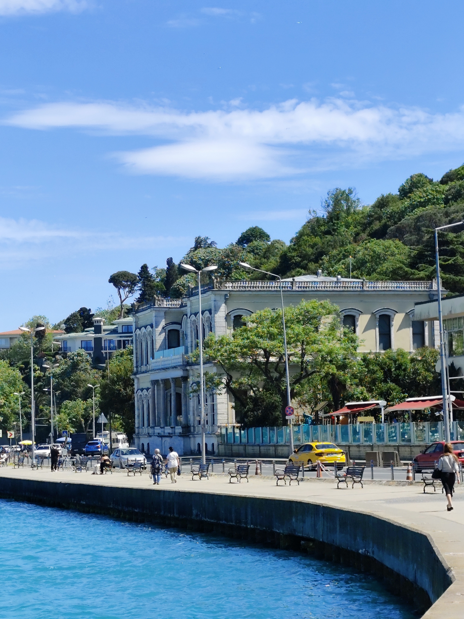 The image depicts a waterfront scene in Sarıyer, Turkey. The Bosphorus Strait is prominent, with its turquoise waters in the foreground. A concrete embankment runs along the water's edge, lined with evenly spaced black benches. People are walking along the pavement.
Behind the pavement is a road with light vehicular traffic, including a yellow taxi. Buildings are on the landside of the road, the architecture featuring ornate facades and structures on the hillside behind. Lush greenery and trees cover the hillsides and grow between the buildings.
The sky is blue with scattered white clouds. Streetlights are visible along the road.