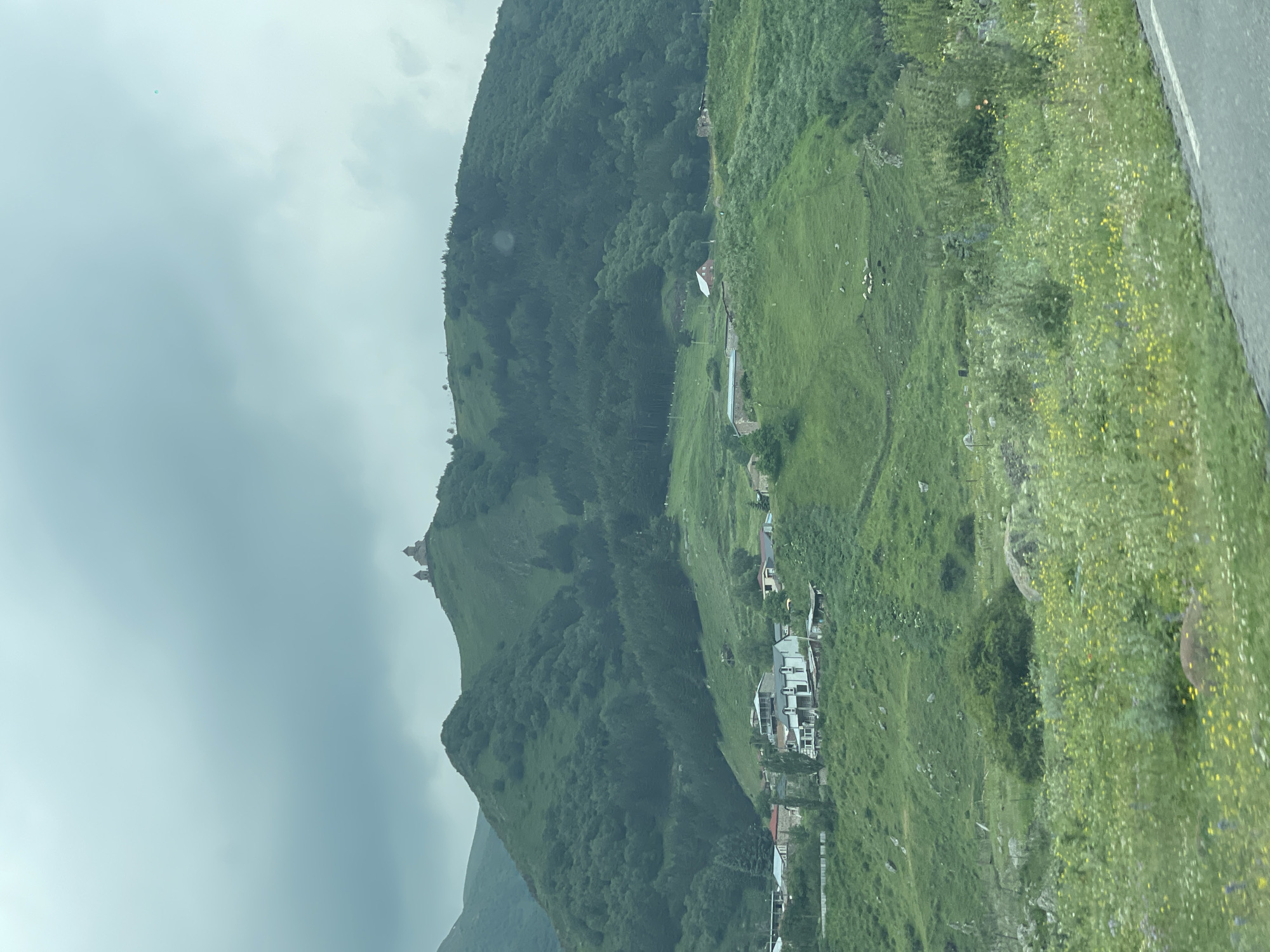 The image shows the Gergeti Trinity Church, a 14th-century Orthodox Christian church located in the city of Gergeti, Georgia. The church is situated on a steep hill, overlooking a valley with a small village below. The landscape is lush and green, with rolling hills covered in trees and wildflowers. The sky is overcast, with dramatic clouds adding to the atmospheric scene. In the foreground, a road is visible, suggesting the vantage point is from a vehicle or a roadside. Several houses and buildings are scattered across the lower slopes of the hills, forming the village. A flock of sheep is grazing in a pasture on the hillside. The overall impression is one of a remote, picturesque, and historically significant location.