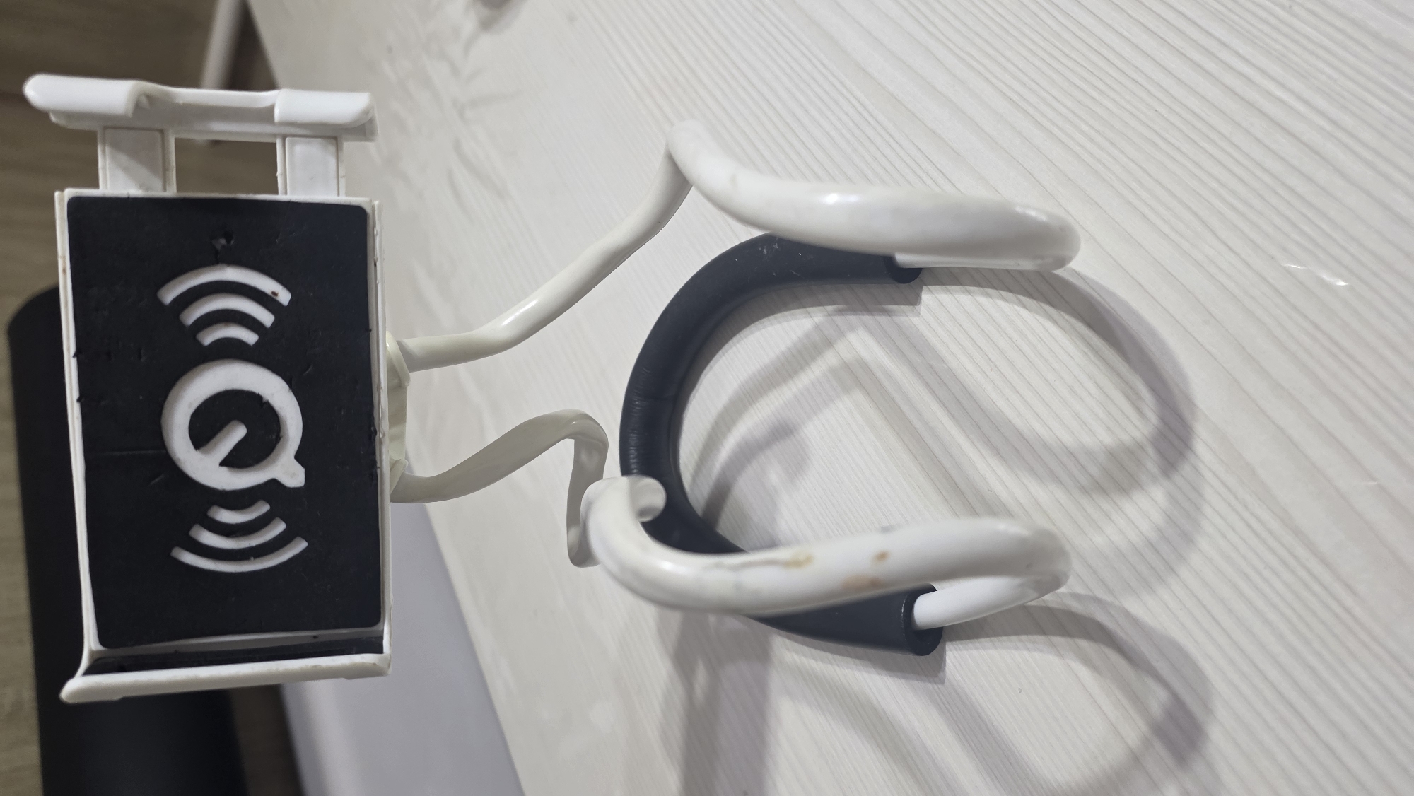 A white and black lazy phone holder is on a white wooden table. The holder has a black rectangular panel with a white logo that appears to be a "Q" with radiating lines on either side. The holder has two white flexible arms that bend downwards, with a black padded section that would rest on a person's neck or chest. The scene is indoors. The location in the city of Chuy, Kyrgyzstan is not identifiable from the image.