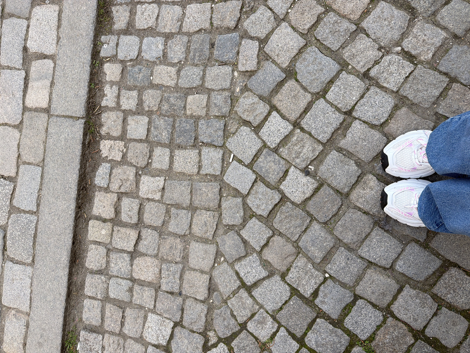 An overhead view captures the lower legs of a person standing on a cobblestone pavement in Sofia, Bulgaria. The individual wears blue denim jeans and white athletic shoes with pink and black details. The cobblestone surface, composed of irregular grey granite blocks, transitions from a diagonal pattern in the foreground to a rectilinear arrangement further back. A dark grey granite curb horizontally bisects the upper portion of the frame, separating the cobblestones from a section of larger, rectangular granite pavers. Sparse green vegetation and minor organic debris are present within the stone gaps and along the curb. Lighting is consistent with diffused daylight.