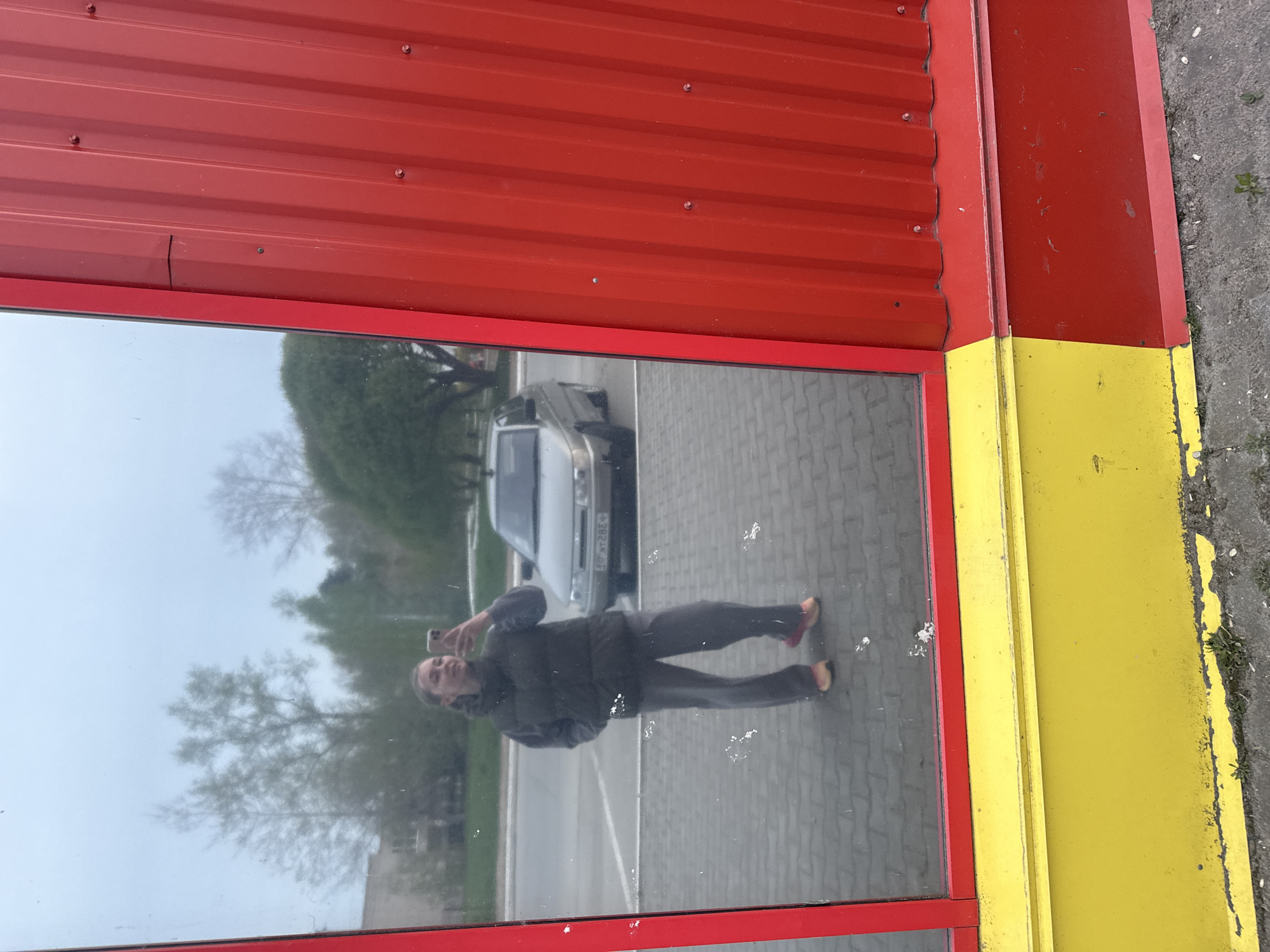 The image shows a reflection in a window. The window frame is red and yellow, and the adjacent wall is red. The reflection shows a person holding a phone, possibly taking a photo of themselves, along with a silver van or car, trees, and pavement. The location is Unknown, Unknown.