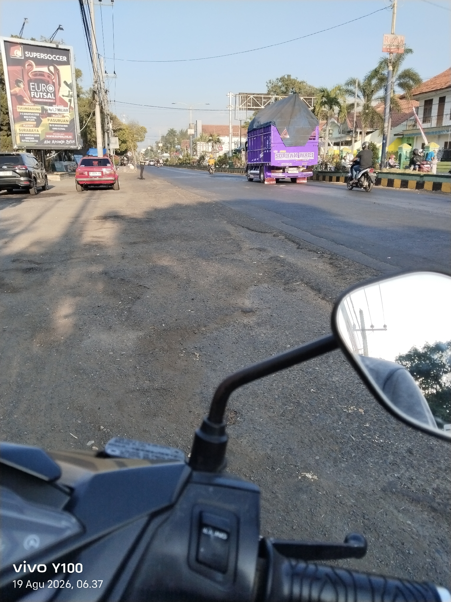 The image captures a view from a motorcycle on a street in Patokan, Indonesia. A billboard for "Supersoccer EURO FUTSAL CHAMPIONSHIP" is prominently displayed on the left side of the road. Various vehicles are present, including a red car, a black SUV, a purple truck with the text "SORBAN WAHYU" on its side, and several motorcycles. People are seen on the motorcycles and walking on the sidewalk. The environment appears to be a mix of urban and semi-urban, with buildings lining the street and palm trees in the background. The sky is clear and blue, suggesting it is daytime. The foreground shows the motorcycle's handlebar and a rearview mirror reflecting the sky and overhead utility poles. The road surface is asphalt with some visible wear and tear. The timestamp on the image indicates it was taken on August 19, 2026, at 06:37.