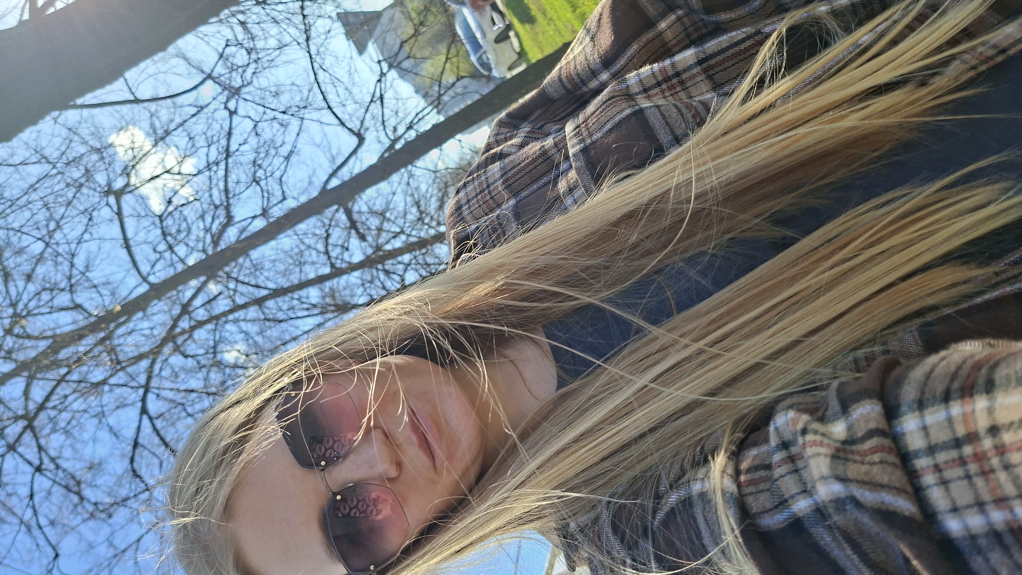A low-angle shot depicts a woman with long, blonde hair wearing sunglasses and a plaid shirt. Bare tree branches against a blue sky are visible in the background, suggesting an outdoor setting. There is a hint of greenery and a vehicle in the distance. The photograph appears to have been taken in Chaikovsky, Russia, based on the location tag.