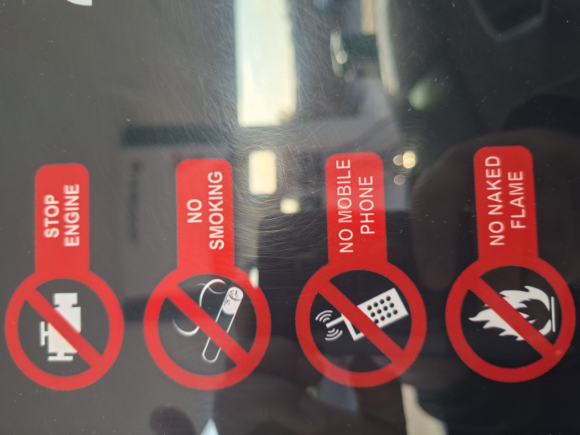 The image displays four red and white prohibition signs, vertically aligned on a dark, scratched, and reflective surface. The background shows blurred reflections of light sources, possibly indicating an outdoor or well-lit indoor setting. These signs are typically found in hazardous environments, such as fuel stations, where safety regulations are critical.
From left to right, the signs convey the following warnings:
1. **STOP ENGINE**: A red rectangular tag with white vertically oriented text "STOP ENGINE". Below it, a white icon of a car engine is depicted within a red circle with a diagonal line, indicating that engines must be turned off.
2. **NO SMOKING**: A red tag with white vertically oriented text "NO SMOKING". Below it, a white lit cigarette icon is crossed out by a red circle and diagonal line, prohibiting smoking.
3. **NO MOBILE PHONE**: A red tag with white vertically oriented text "NO MOBILE PHONE". Below it, a white mobile phone icon showing radiating signals is prohibited by a red circle and diagonal line, indicating that mobile phone use is not allowed.
4. **NO NAKED FLAME**: A red tag with white vertically oriented text "NO NAKED FLAME". Below it, a white stylized flame icon is crossed out by a red circle and diagonal line, prohibiting any open flames.
No people are visible in the image. There are no specific cues to determine the exact time of day or weather conditions. The signs are generic safety warnings, located in Plovdiv, Bulgaria, as per the provided context.