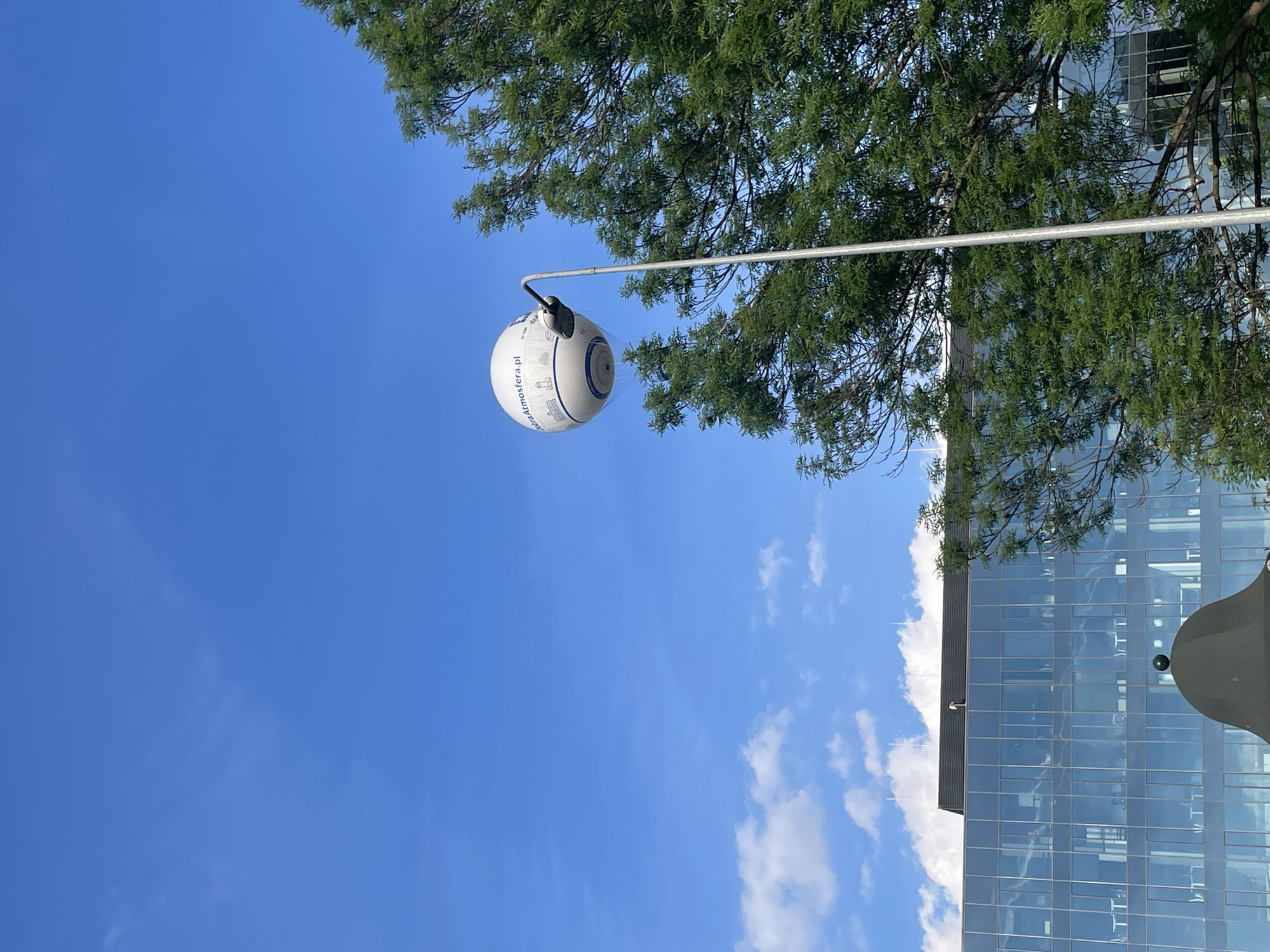 The image shows a white sphere attached to a metal pole, suspended in the air against a backdrop of blue sky with a few clouds. The sphere has a circular design with blue lines and text that appears to read "atmosfera.pl". Green foliage from a tree overhangs the sphere and the metal pole. A tall, modern building with glass windows is partially visible on the right side of the image. The location is potentially somewhere in the city of Unknown, Unknown.
