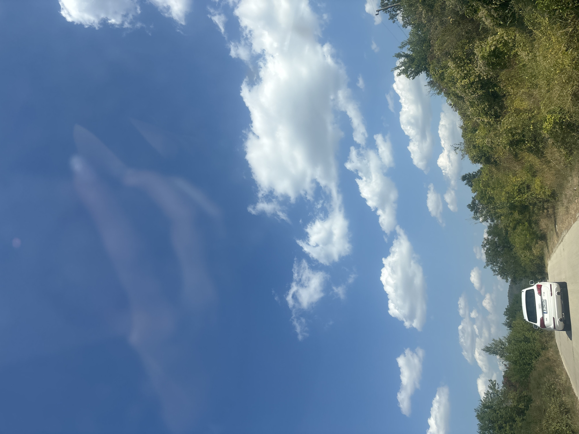 A white Audi Q5 SUV is driving on a paved road through a rural, wooded area. The sky is bright blue with scattered white clouds, and there are some blurry, light-colored reflections in the upper portion of the sky that suggest the image was taken from inside a vehicle. The road appears to be a single lane in each direction, with a grassy shoulder and dense green vegetation on either side. The trees are a mix of deciduous and evergreen species, and the vegetation is lush and overgrown. The overall impression is of a warm, sunny day in the countryside. The location is not specified in the image.