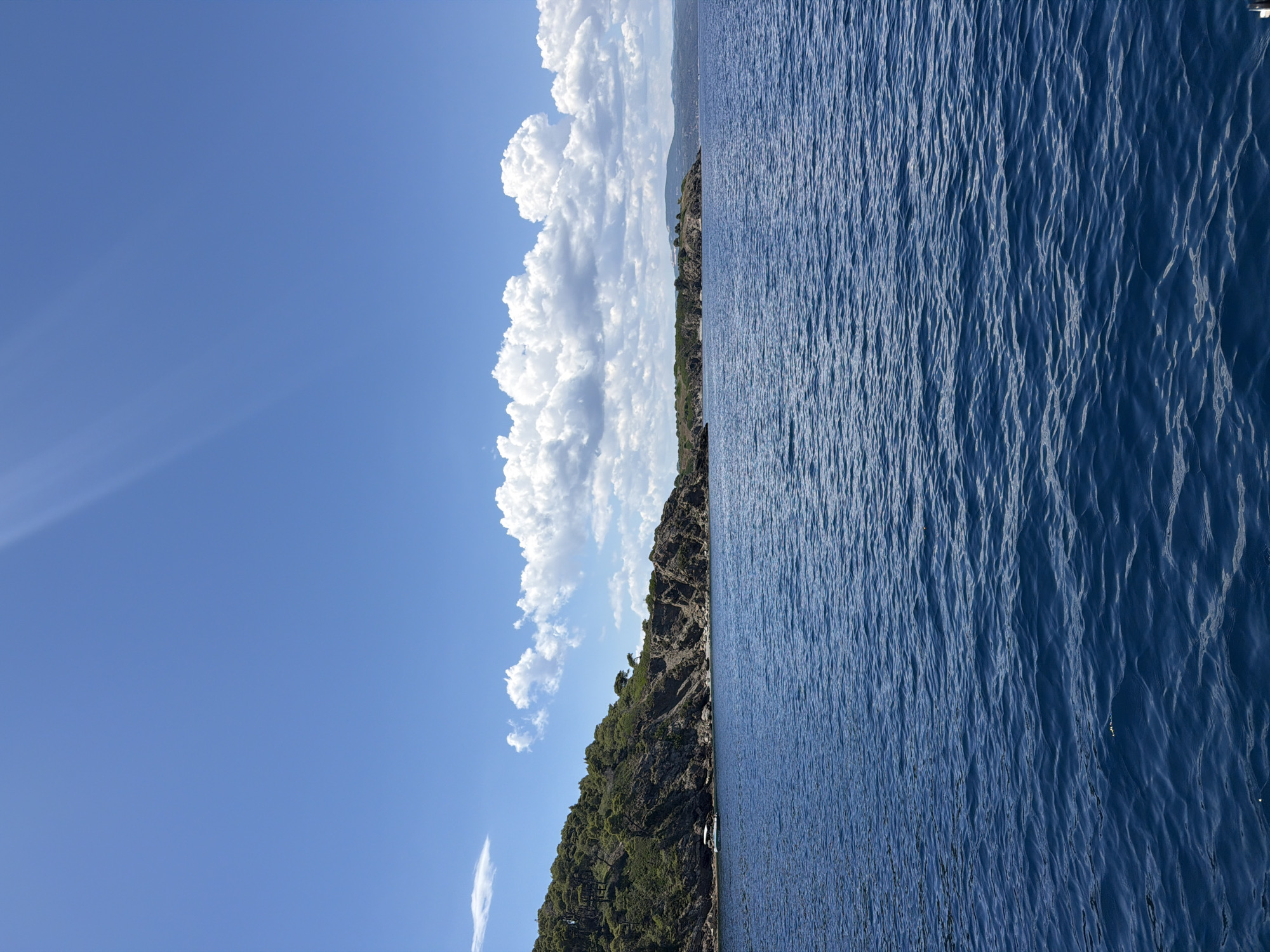 The image depicts a scenic view of the Aegean Sea off the coast of an island in Greece. The foreground is dominated by the deep blue, rippling water of the sea. In the mid-ground, a lush green, rocky island juts out into the water. The island is covered with dense vegetation, primarily trees and shrubs, with rocky outcrops visible along its shoreline and higher elevations. A small, white boat with an awning is anchored near the shore of the island. The background features a clear, bright blue sky with scattered white, fluffy cumulus clouds. Sunlight streams down from the upper left corner of the frame, casting a slight glare. The distant horizon shows a faint outline of more landmass, possibly other islands or the mainland, under the expansive sky. The overall atmosphere is serene and indicative of a sunny day at sea. The specific location is not identifiable from the image alone, but the landscape is characteristic of the Greek islands.