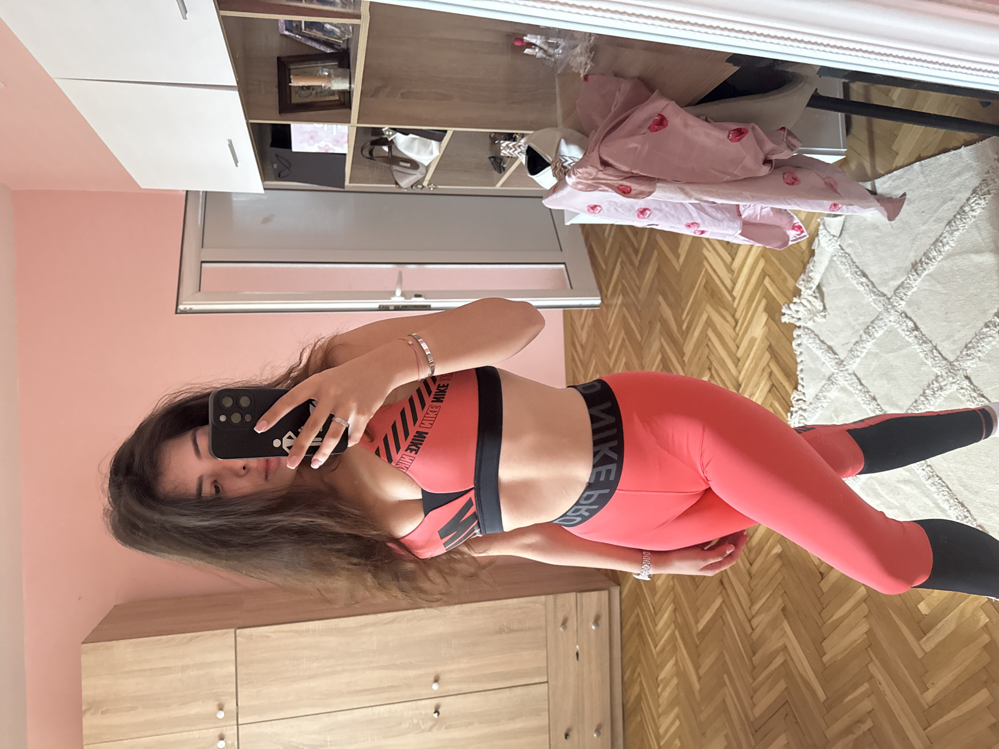 A young woman with long, dark, wavy hair stands in a room, taking a mirror selfie. She is wearing a bright coral-colored sports bra and matching leggings with black accents. The sports bra has "NIKE" written across it in black text. The waistband of the leggings also features "NIKE" branding. She has on a silver wristwatch and a delicate silver bracelet on her left wrist, and a ring on her right index finger.
The room has pale pink walls. To the left of the woman is a tall, light wood wardrobe with two doors. The floor is covered in a light-colored parquet flooring arranged in a herringbone pattern. In the background, to the right, a white wardrobe with shelving is visible, containing various items including framed photos and bags. A partially visible doorway with a white door is also present. A rug with a diamond pattern and tasseled edges lies on the floor in front of the woman. A pink garment with a pattern of small red hearts is draped over a dark-colored chair or stand near the white wardrobe.
The overall scene suggests a domestic setting, possibly a bedroom or dressing area, in a residential apartment or house located in the city of Plovdiv, Bulgaria. The lighting appears to be natural, coming from a light source not directly visible in the frame.
