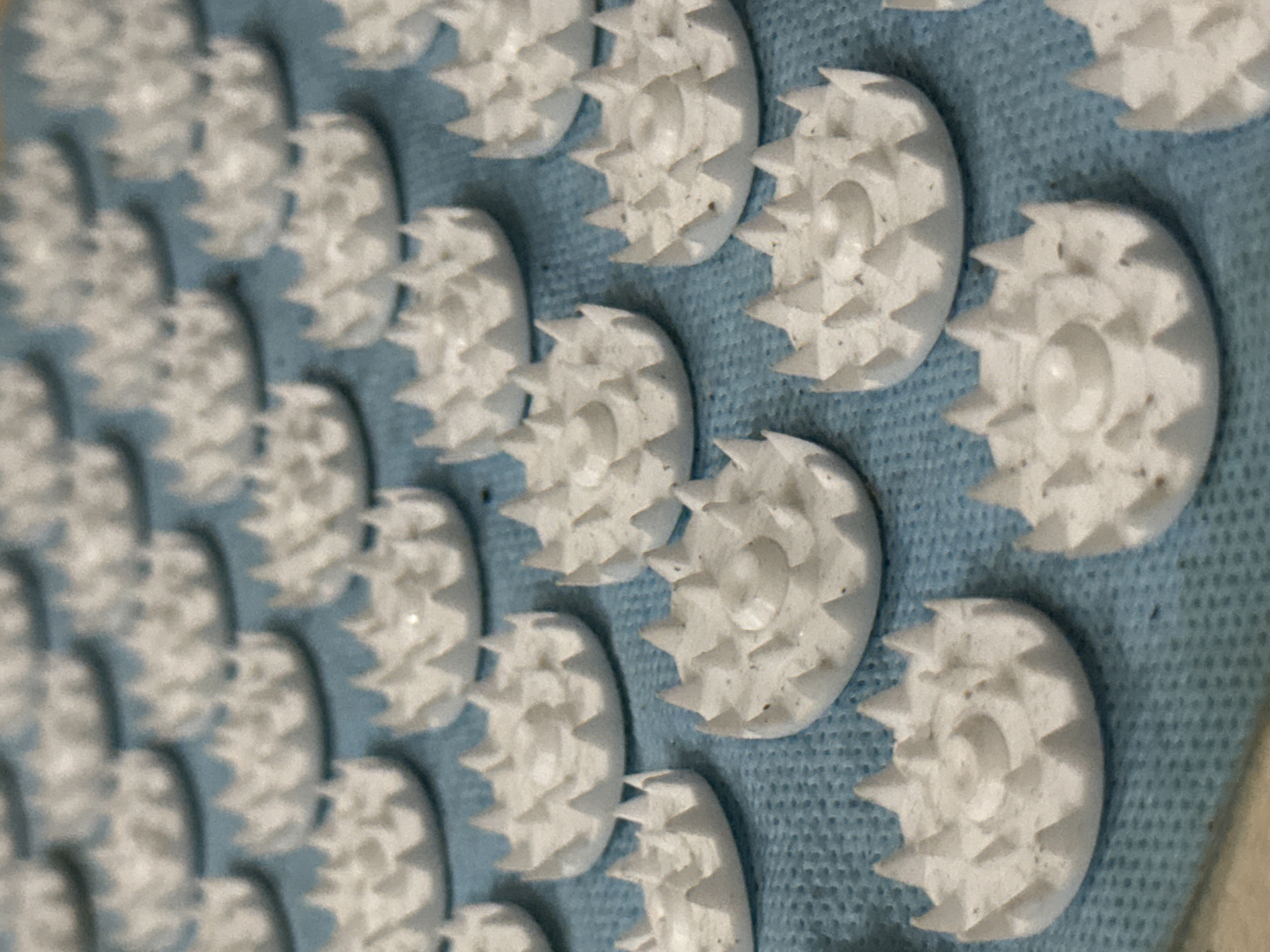 A close-up shot presents an array of white, circular objects with spiky edges arranged on a light blue surface. The circular objects have a small, raised center and multiple triangular projections extending outward. The arrangement is somewhat grid-like, with objects positioned closely together. The background surface appears to be textured fabric. The image suggests a manufactured item, potentially a component of a massage or acupressure product. The location of the city of Đà Nẵng, Vietnam is unknown.