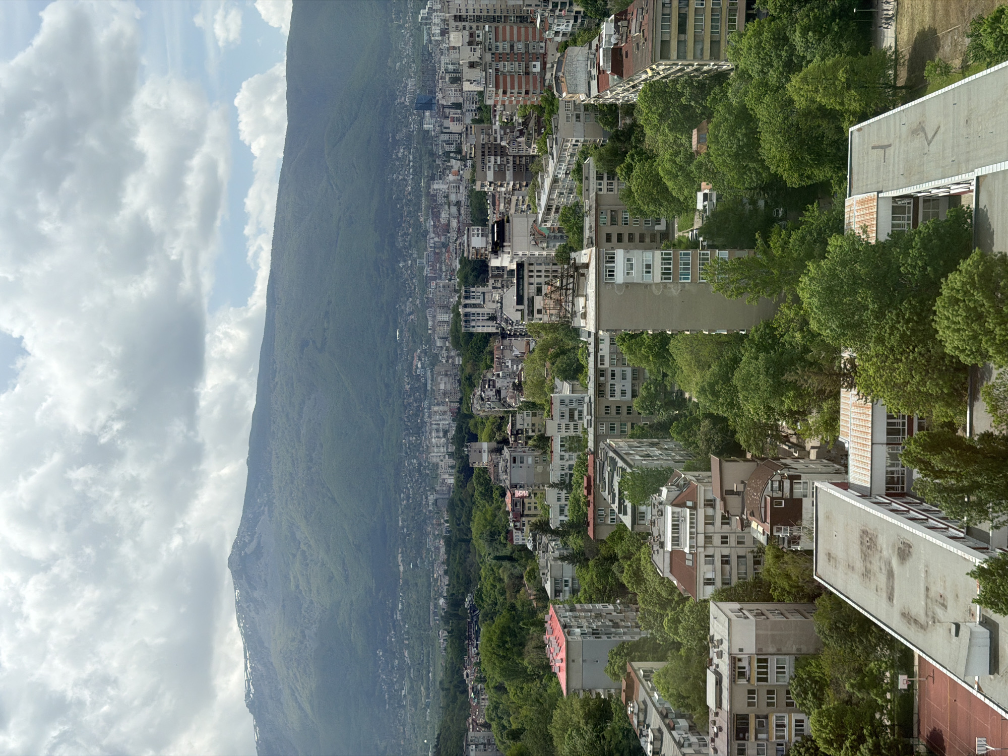 The image shows a view of the city of Sofia, Bulgaria, with the Vitosha Mountain in the background. The foreground and middle ground are populated with buildings of varying heights, interspersed with green trees. The mountain in the background is a large, green landmass that slopes down towards the city. The sky is cloudy. The image appears to have been taken from a high vantage point.