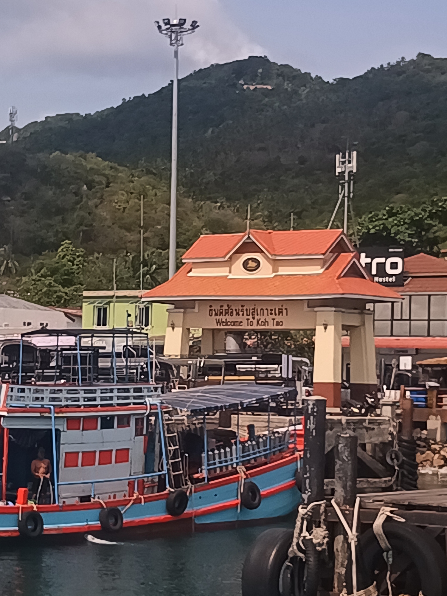 The image shows a pier in Tambon Ko Tao, Thailand. In the foreground, a brightly colored blue and red boat is docked. A person is visible onboard the boat. The pier itself is constructed of wooden posts and tires are used as bumpers. In the background, there is a welcome sign with a red roof and cream-colored pillars that reads "Welcome to Koh Tao". Behind the sign is a building with a sign that reads "Retro Hostel". The background features a mountainous landscape covered in green vegetation. A tall light post and cell tower are present on the mountainside. The sky is overcast.