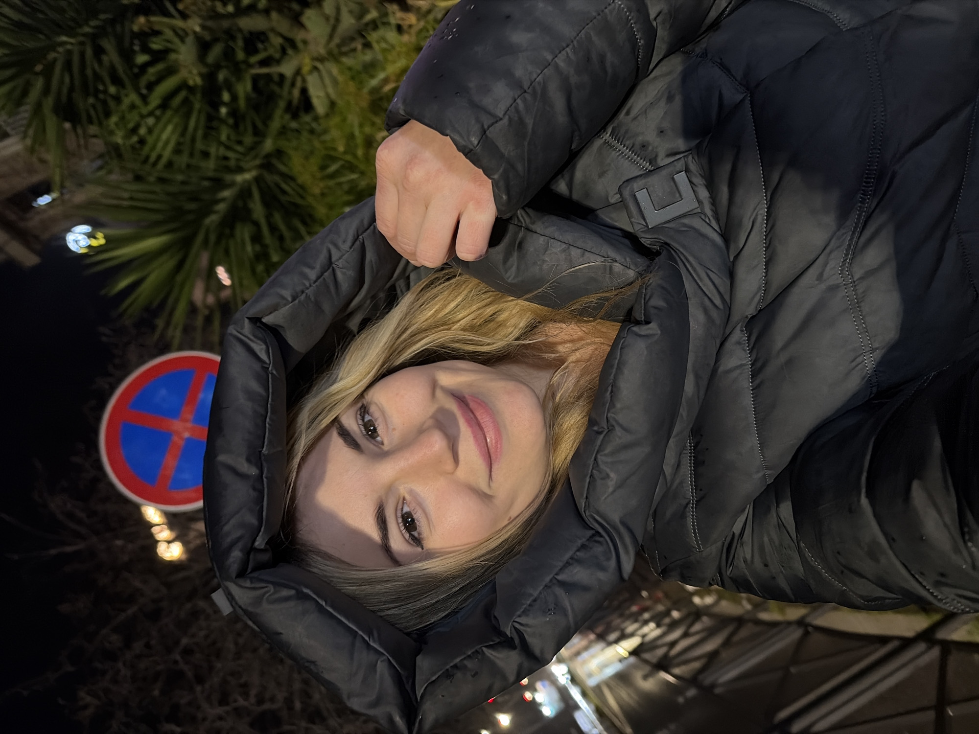A woman with blonde hair, wearing a black puffer jacket with the hood up, is in the frame. She is smiling and looking at the camera. Behind her is a "no parking" sign with a red cross on a blue circle. The background appears to be a street scene at night, with street lights and some greenery visible. Based on the "no parking" sign design and general appearance, the location is likely Budva, Montenegro.