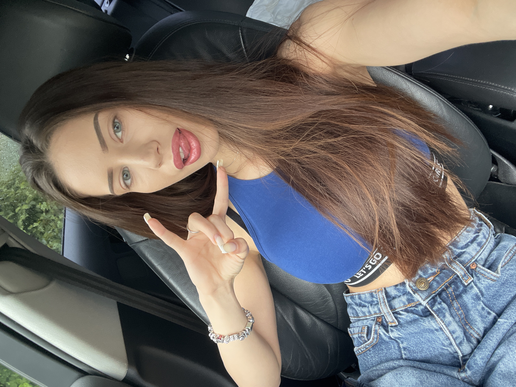 A young woman with long, brown hair and blue eyes is sitting in the driver's seat of a car. She is wearing a blue crop top and denim jeans, and she is sticking her tongue out and making a peace sign with her fingers. The car's interior is visible, including the black leather seats and a seatbelt. The background is blurred, but appears to be foliage.