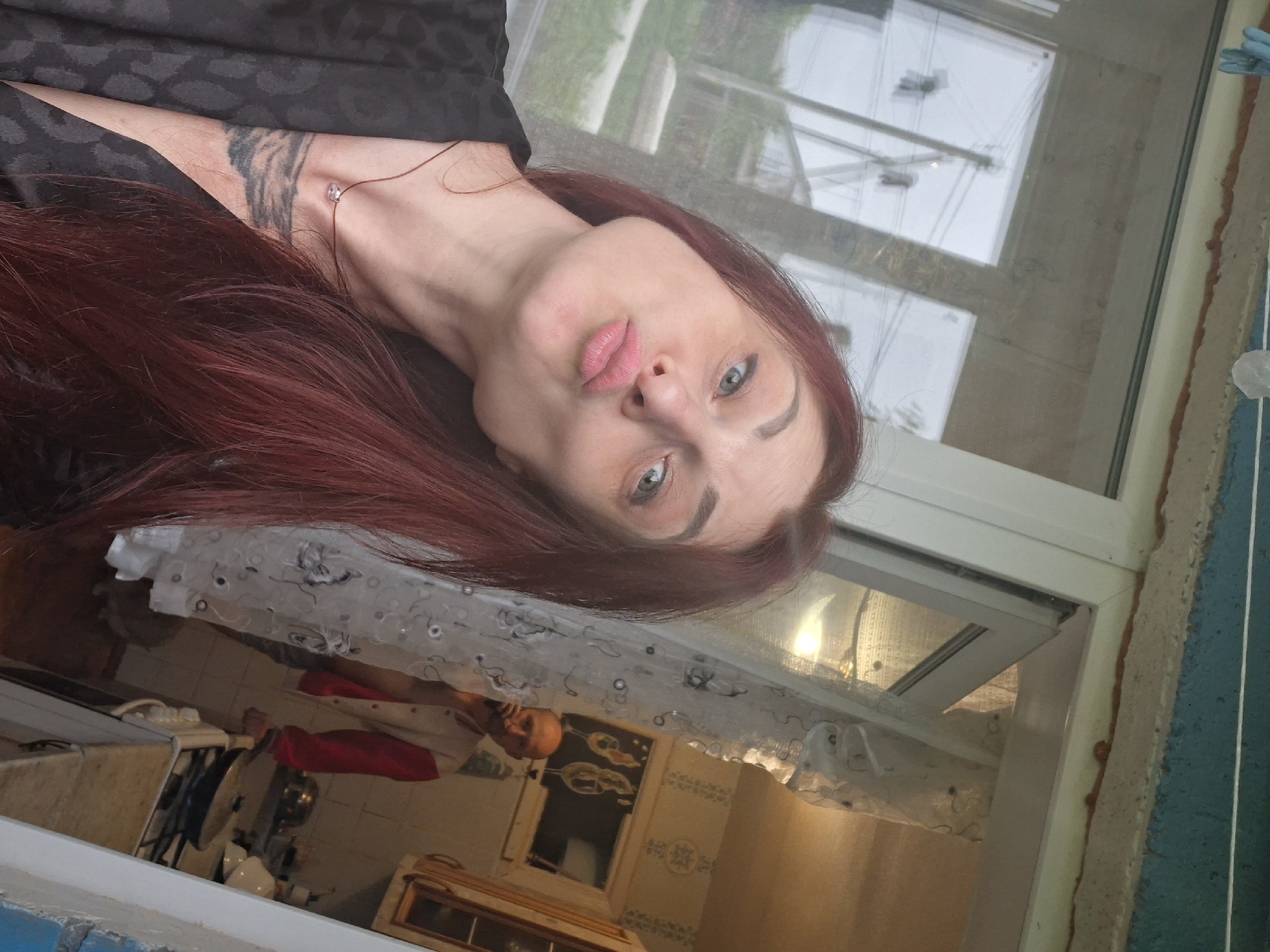 A woman with long, red hair and fair skin is taking a selfie, puckering her lips. She is wearing a black patterned robe and a thin necklace with a small pendant. A tattoo is visible on her left forearm. In the background, a man with a shaved head is standing in a kitchen, looking at his phone. The kitchen appears to be cluttered, with cabinets and a stove visible. The scene is lit by natural light coming from a window or door. The location is identified as Забалоцкі сельскі Савет, Belarus.