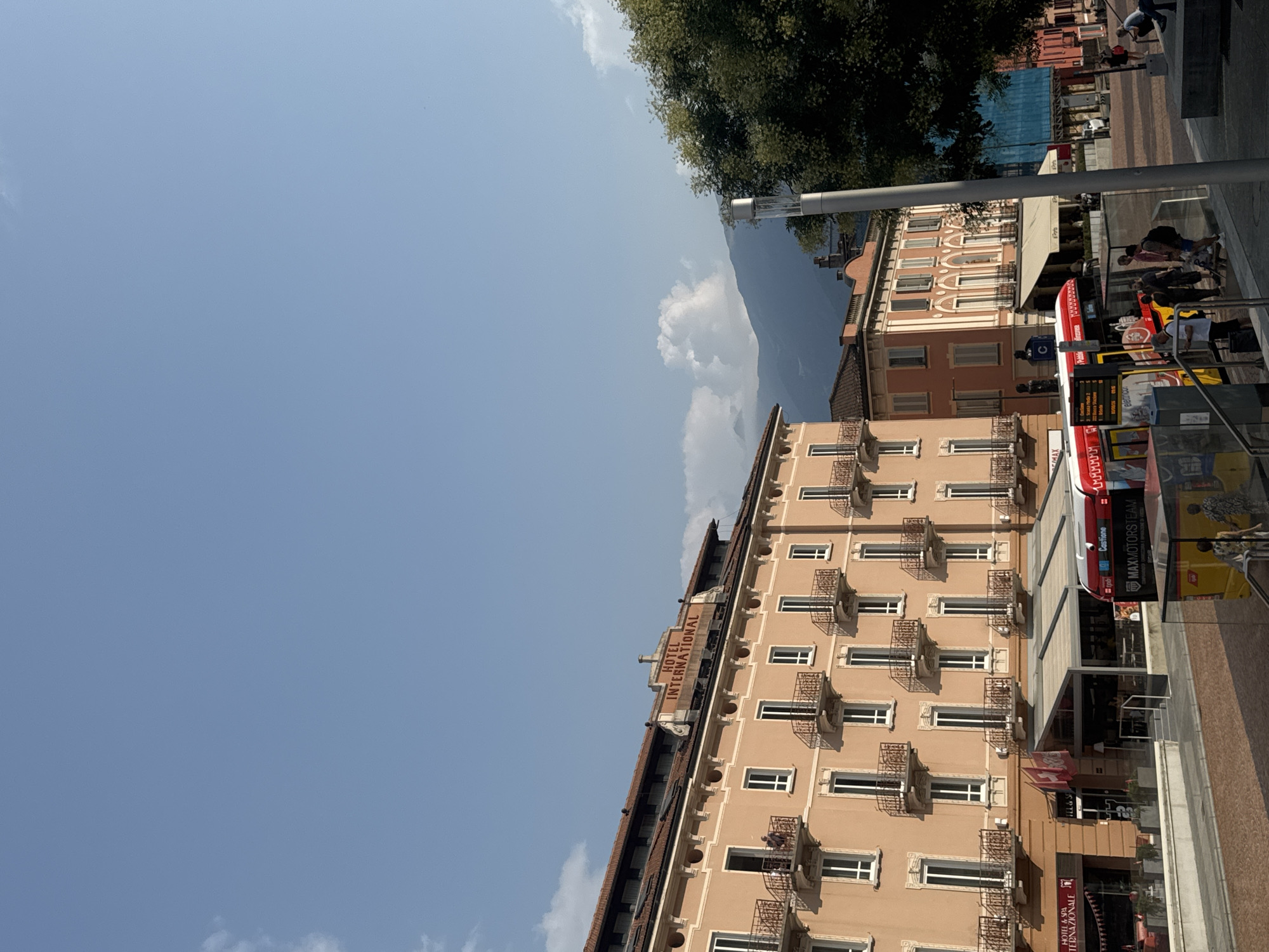The image captures a view of the Hotel Internazionale in Bellinzona, Switzerland, on a clear day with a few clouds. The hotel, an ochre-colored building with multiple floors, features ornate balconies with black wrought-iron railings. The name "HOTEL INTERNATIONAL" is prominently displayed in red lettering above the main facade.
In the foreground, a red and yellow public transportation bus is visible, indicating a busy urban setting. People are seen standing near the bus and on the sidewalk, some appearing to be waiting or disembarking. The street in front of the hotel is paved with cobblestones.
Behind the hotel, a range of mountains is visible, softened by the distance and a slight haze. A tall streetlamp stands to the right of the frame, partially obscuring a lush green tree. Other buildings, including a pink and orange structure with arched windows, are also present, contributing to the cityscape. The overall scene suggests a vibrant and active town square or street.