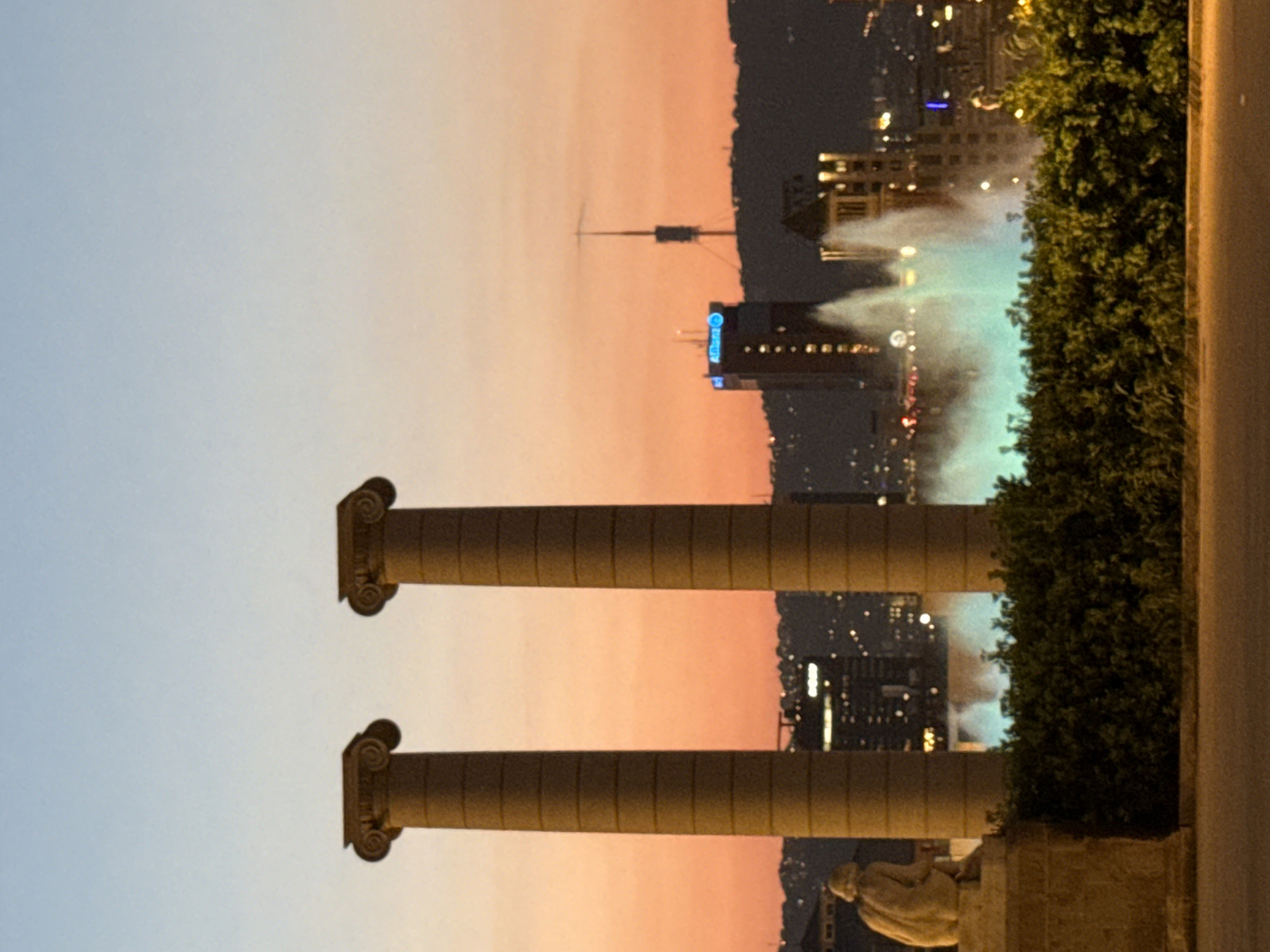 The image depicts a panoramic view of Barcelona at dusk. In the foreground, two tall, fluted columns with Corinthian capitals stand prominently. To the left of the columns, a stone statue of a seated figure is visible on a pedestal. Behind these architectural elements, a city skyline emerges against a gradient sky of soft blues and warm oranges. The city is illuminated with scattered lights, indicating nighttime activity. A notable landmark in the background is a tall tower with an antenna structure. In the mid-ground, a dynamic fountain shoots jets of water, illuminated with a turquoise glow, into the air. Dense green foliage lines the area in front of the fountain and the city. The overall scene captures a blend of ancient architecture, modern cityscape, and natural elements under a twilight sky. The location is likely Montjuïc hill in Barcelona, given the presence of the Magic Fountain of Montjuïc and the characteristic skyline.